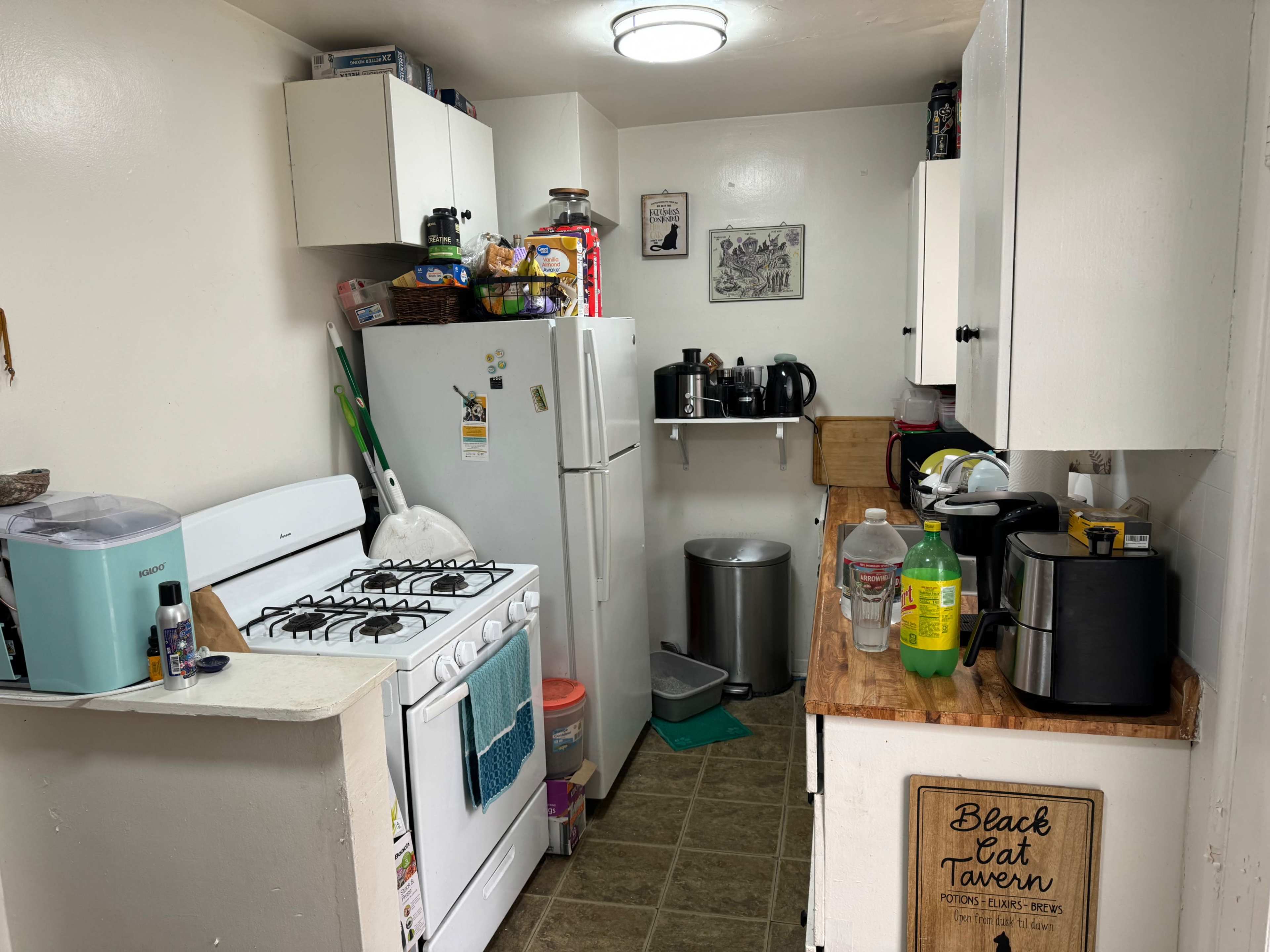 The image shows a small kitchen with white cabinets, a gas stove, a refrigerator, and various appliances arranged on a wooden countertop.
