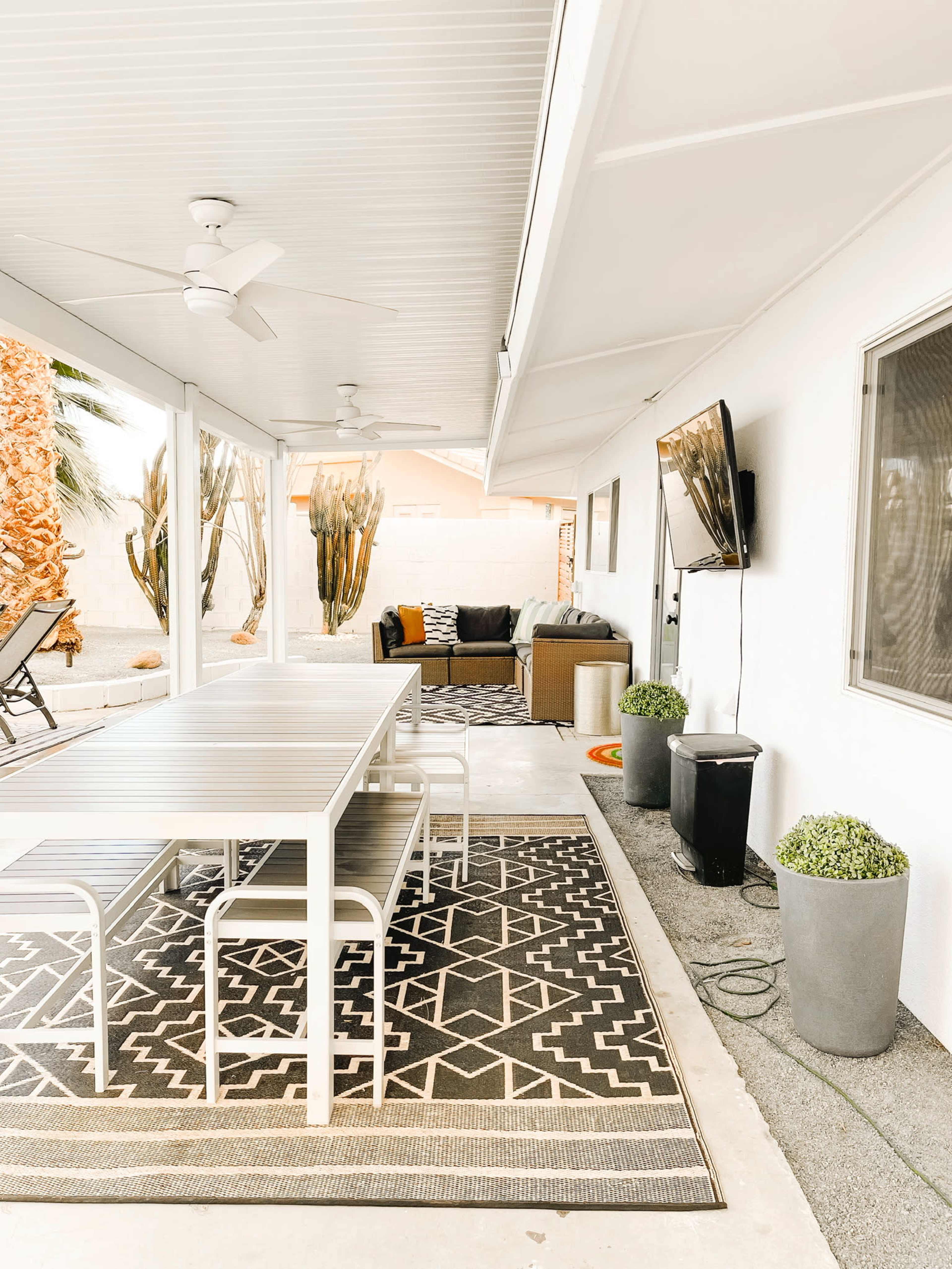 The image shows a covered outdoor patio with a dining table, comfortable seating area, and potted plants, all set against a backdrop of palm trees.