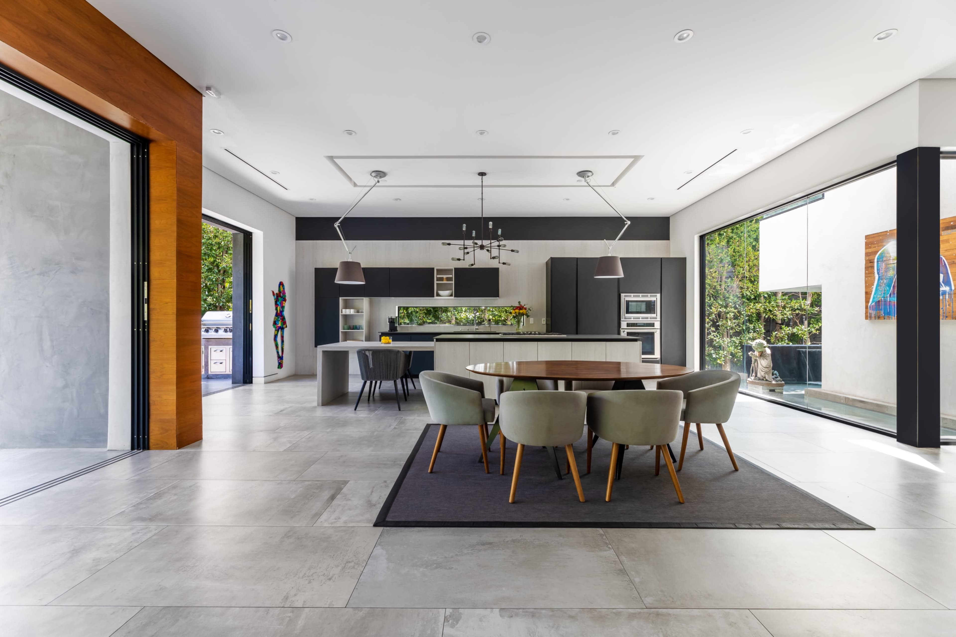 The image depicts a modern kitchen and dining area with a large table surrounded by upholstered chairs, large windows, and sleek cabinetry.