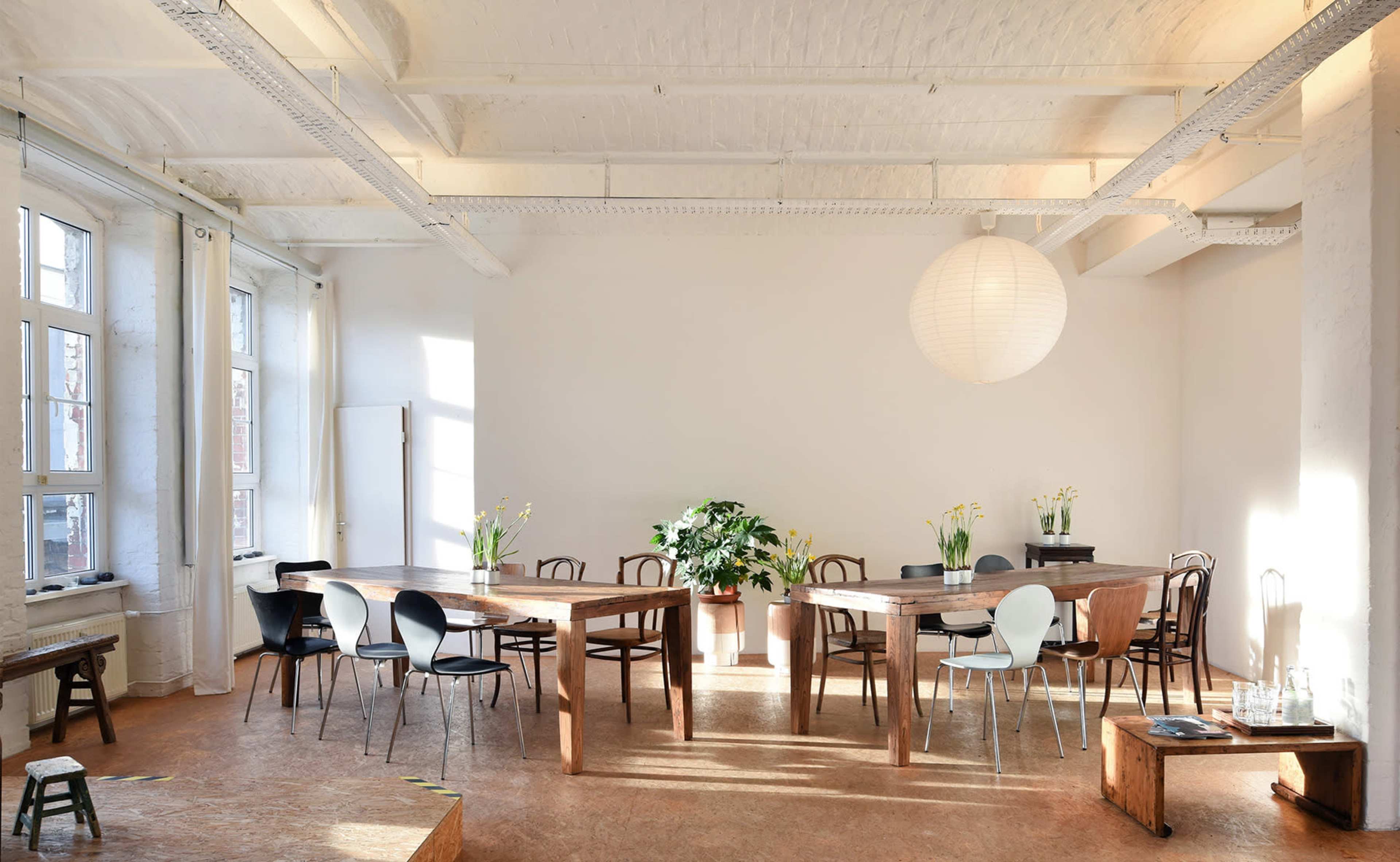A spacious, well-lit room features a large wooden table surrounded by various chairs and potted plants.