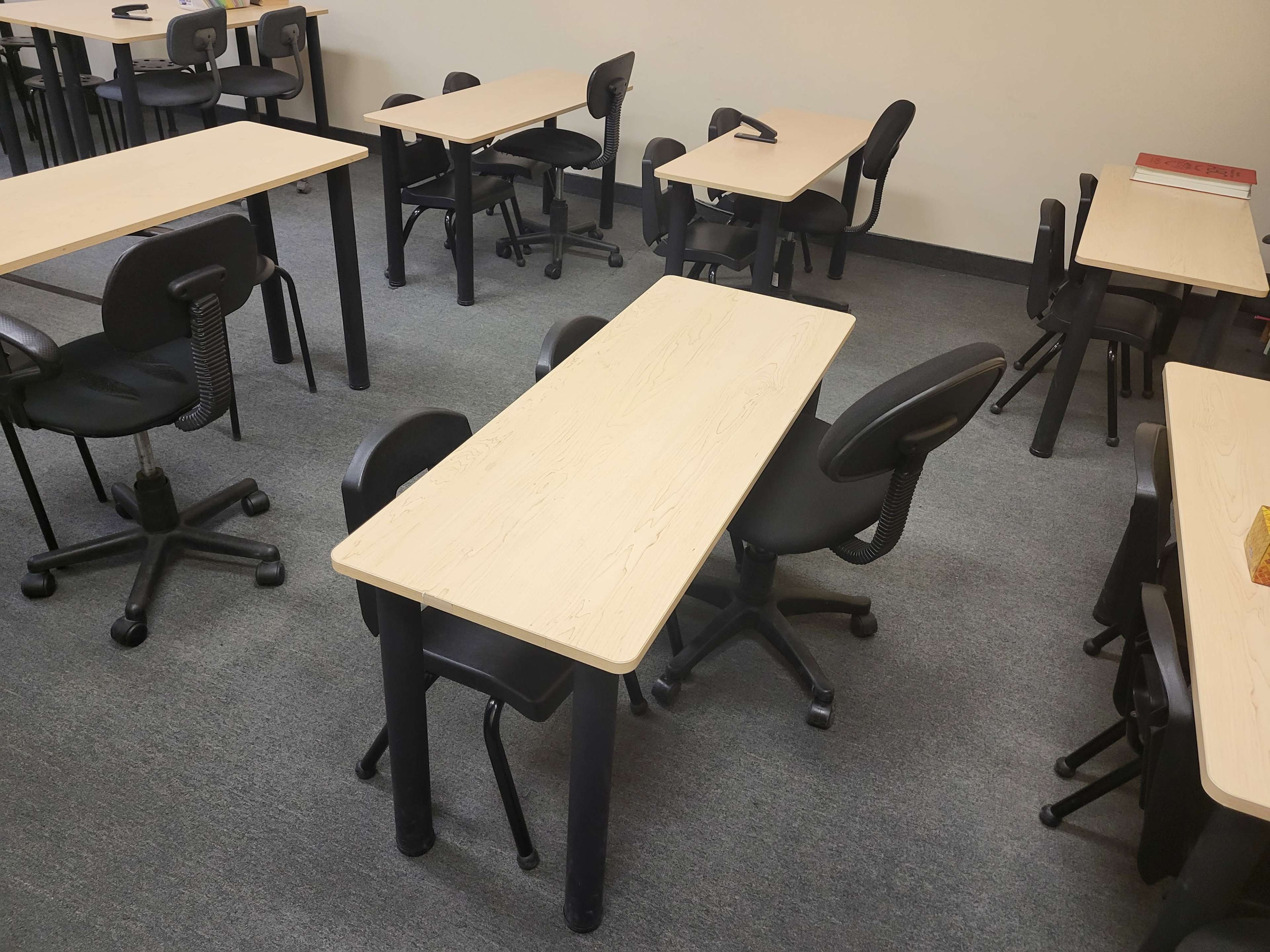 A classroom with several wooden tables and black rolling chairs arranged on a gray carpet.