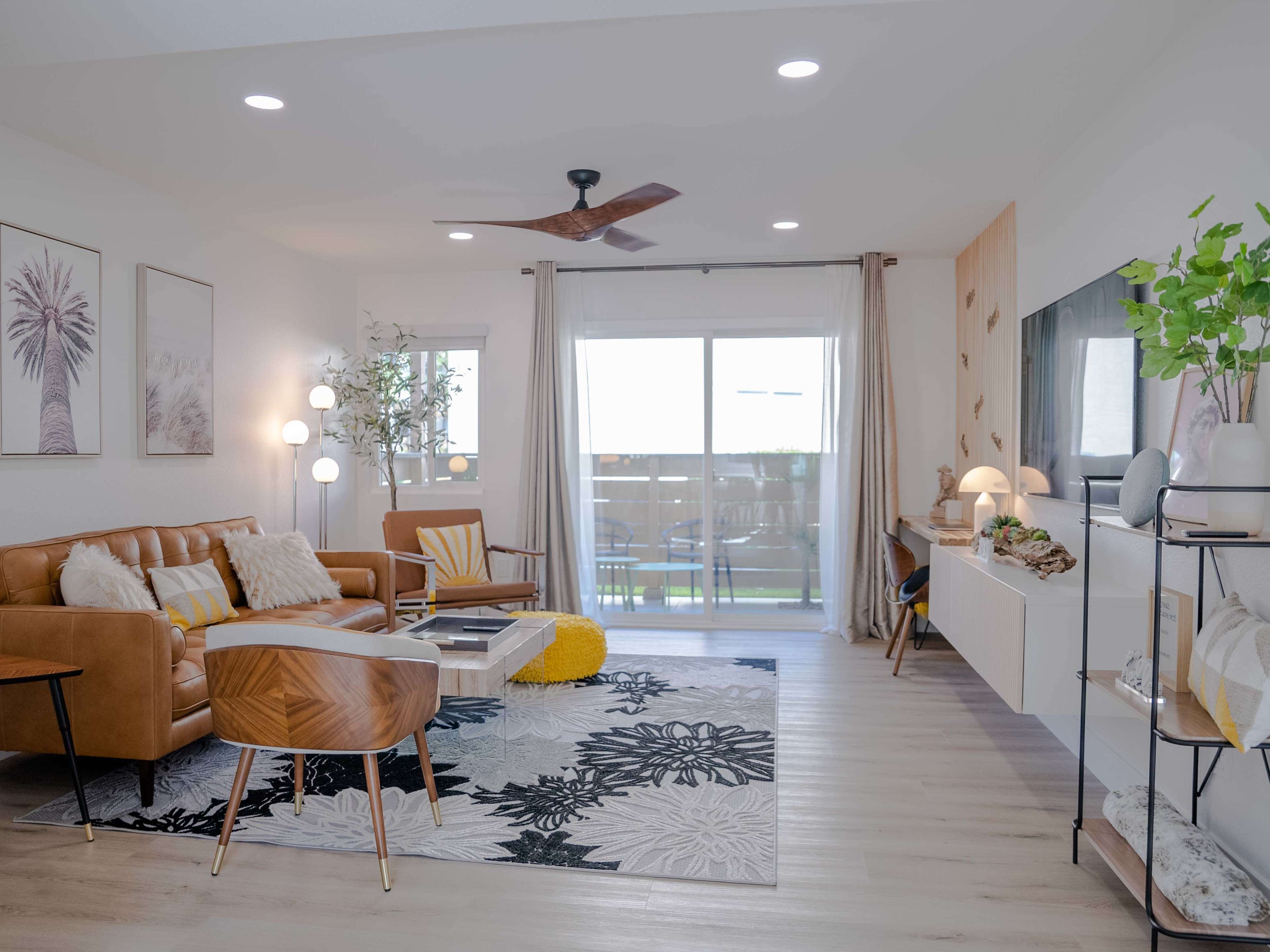 A modern living room features a brown leather sofa, a glass coffee table, and a large window with sliding doors, complemented by a decorative rug and plants.