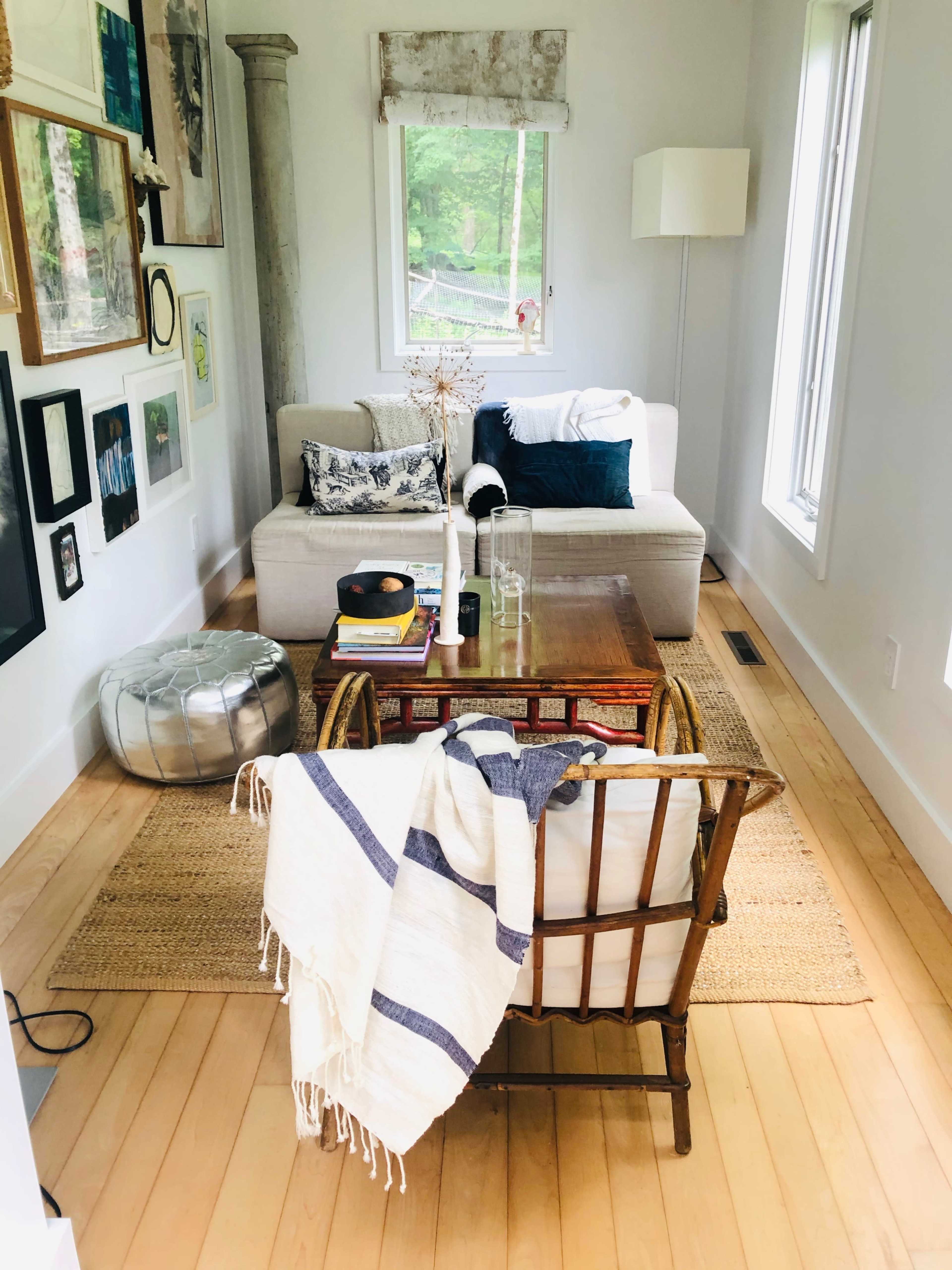 The image shows a cozy living room featuring a light-colored sofa, a glass coffee table, a patterned throw rug, and a chair with a striped blanket, surrounded by numerous framed artworks on the walls.