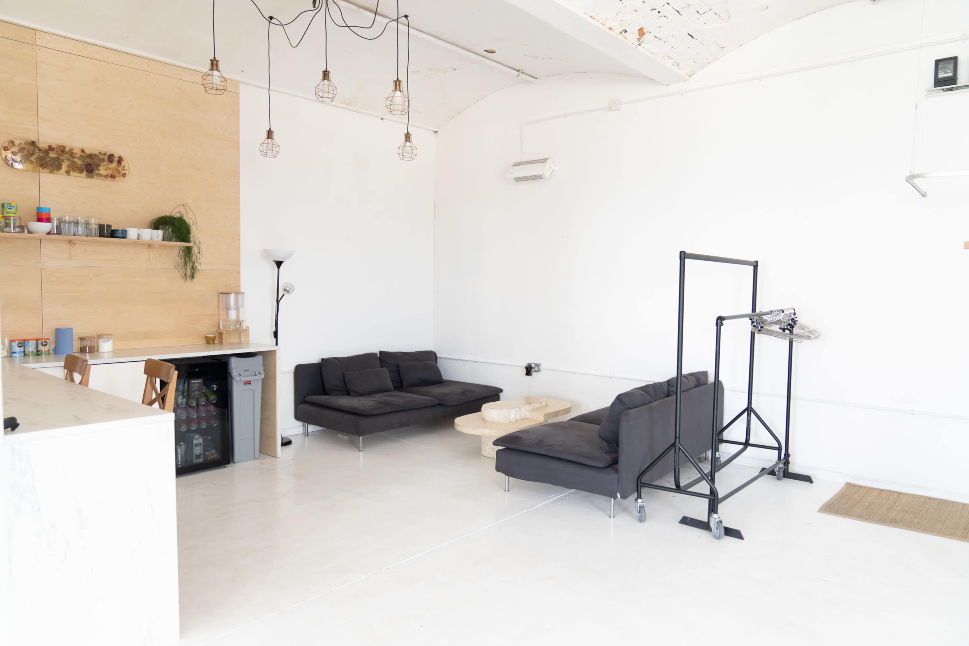The image shows a minimalist interior featuring two dark sofas, a coffee table, a small kitchenette area, and a clothing rack in a white space with wooden accents.
