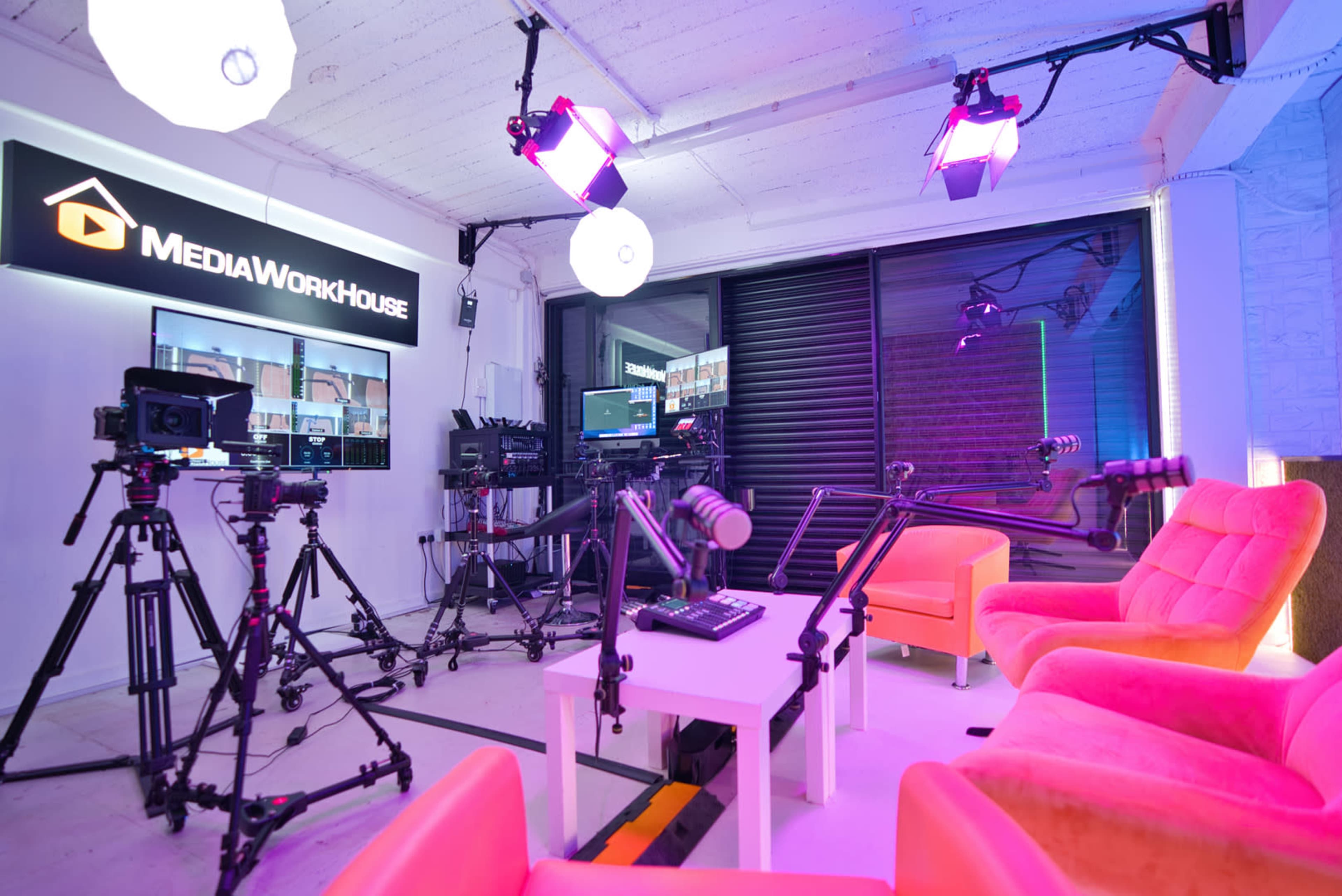 A brightly lit media studio with several cameras, microphones, and two pink chairs arranged around a small table.