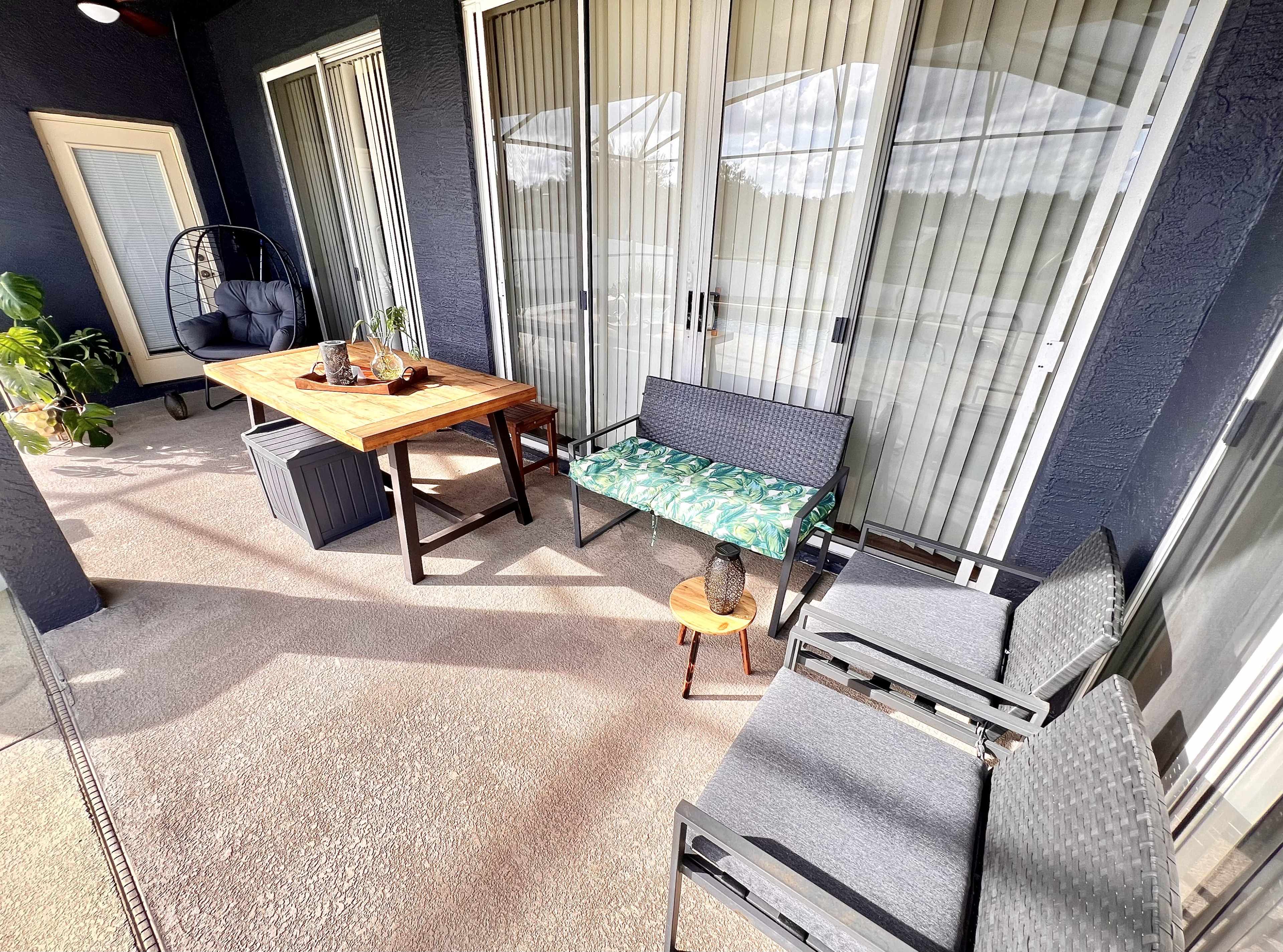 The image shows a patio area featuring a wooden table, a bench with a green cushion, and multiple seating options, all surrounded by large glass doors.