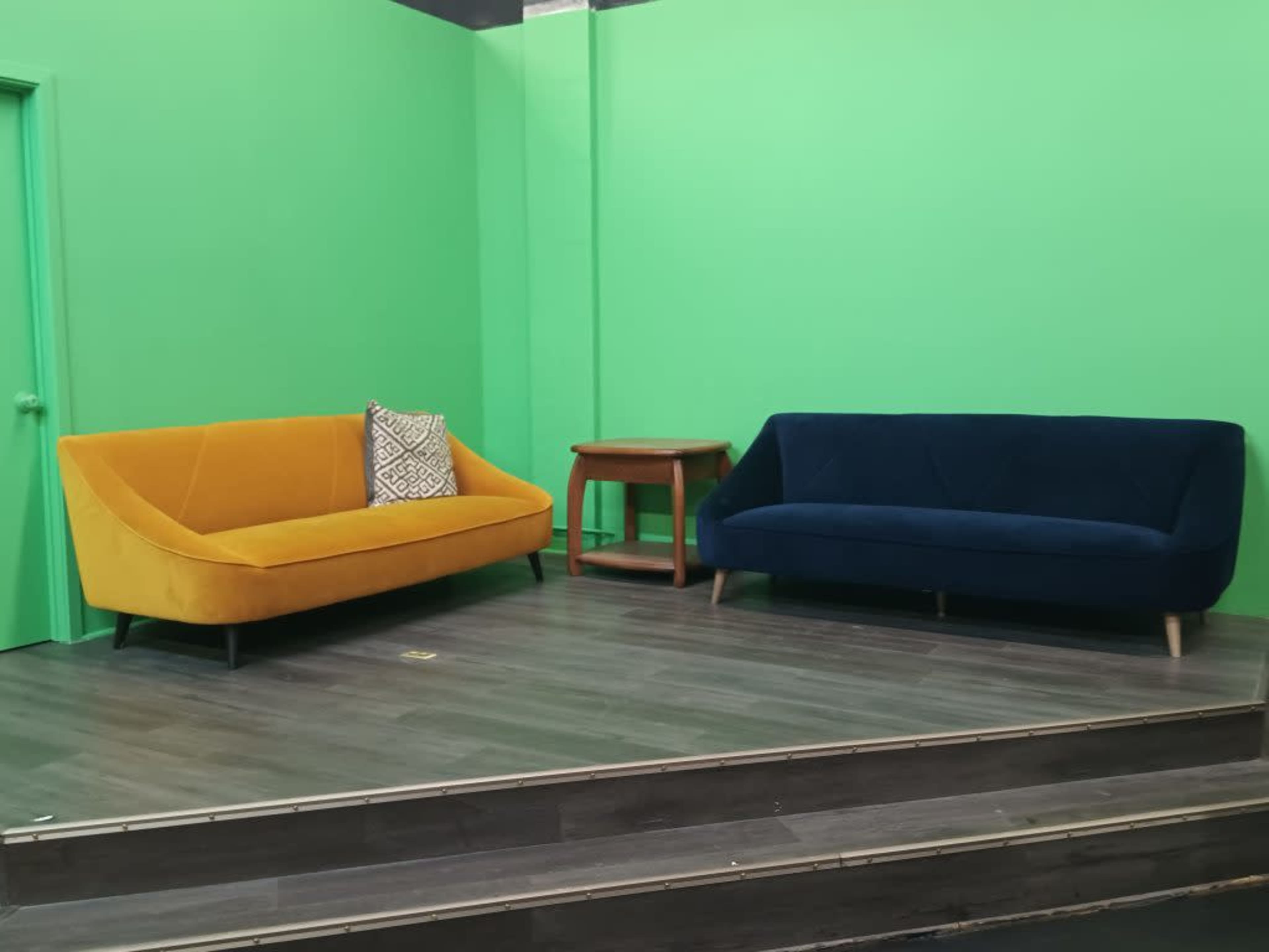 A yellow couch and a blue couch are positioned on a wooden floor in front of a green screen backdrop.