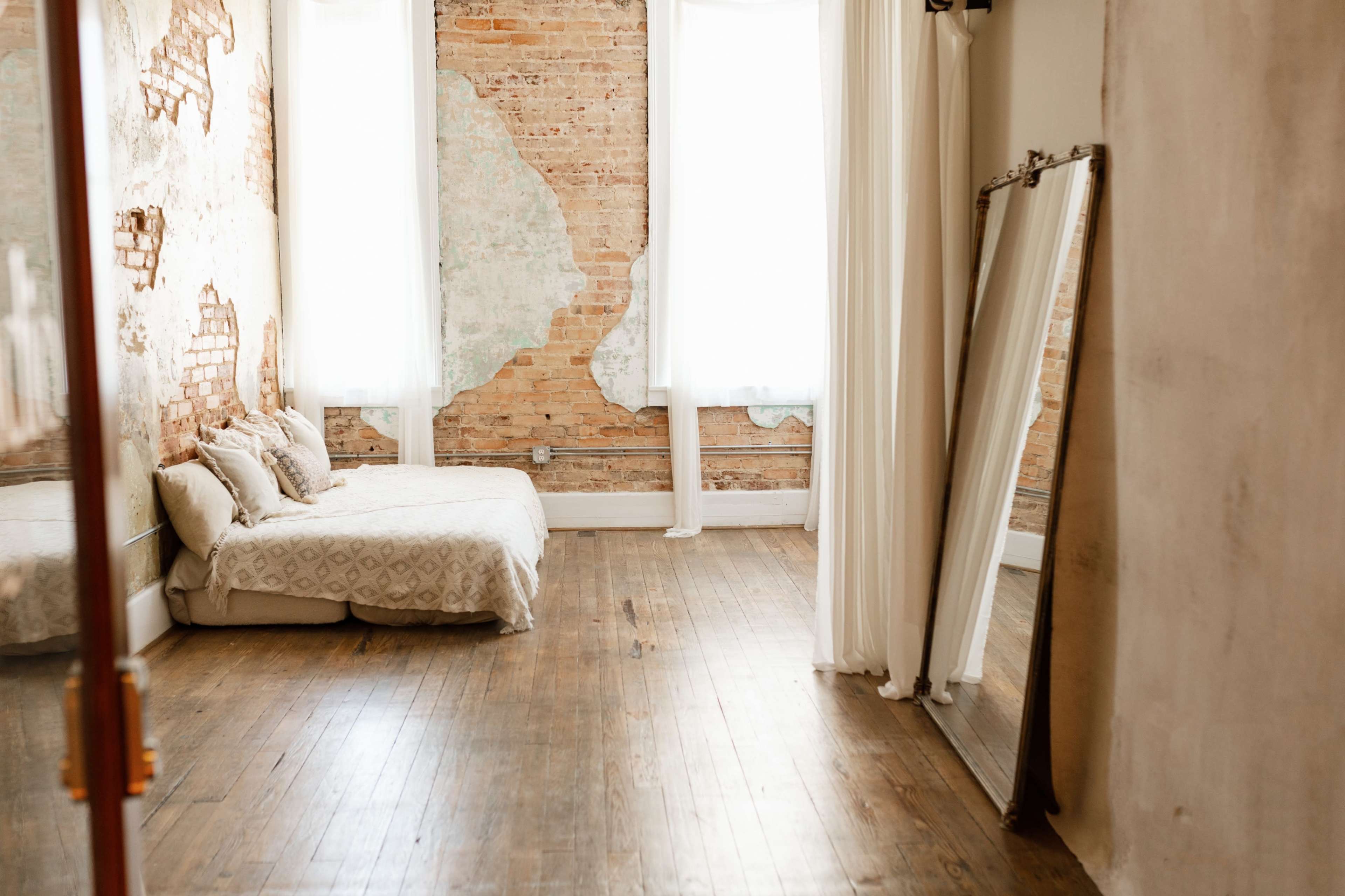 The image shows a minimalist bedroom with a bed on a wooden floor, exposed brick walls, and large windows allowing natural light to enter.
