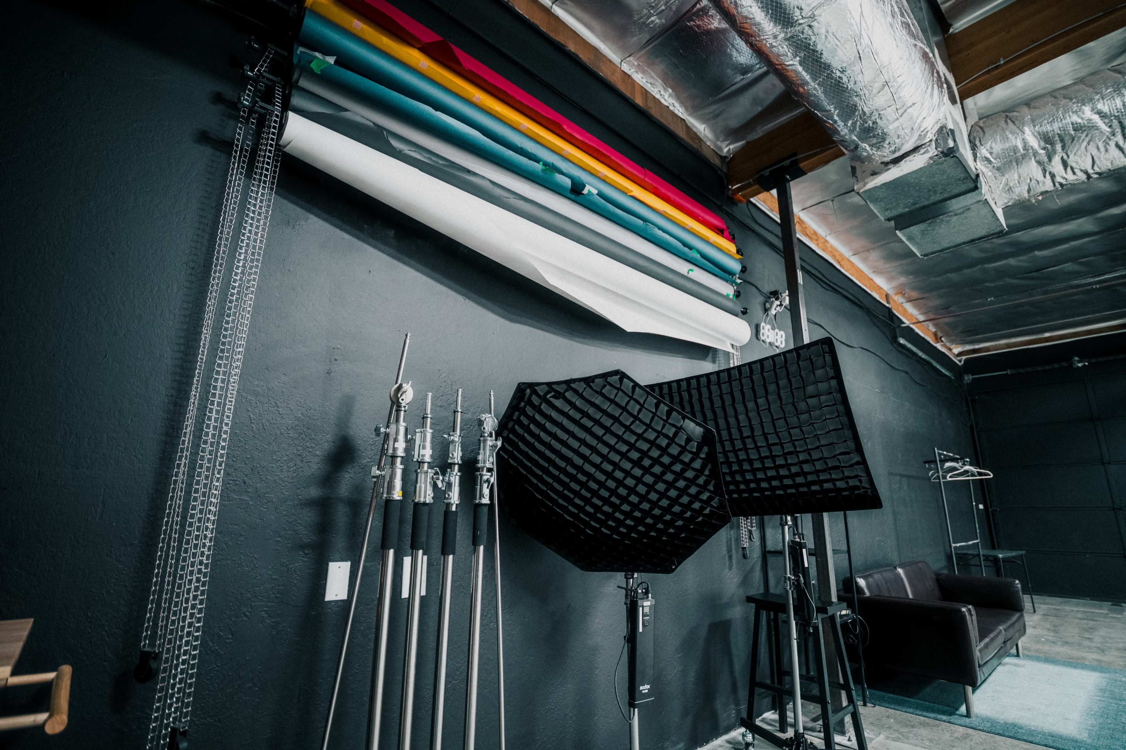 The image shows a photography studio with rolled-backdrops, lighting equipment, and a black couch against a dark wall.