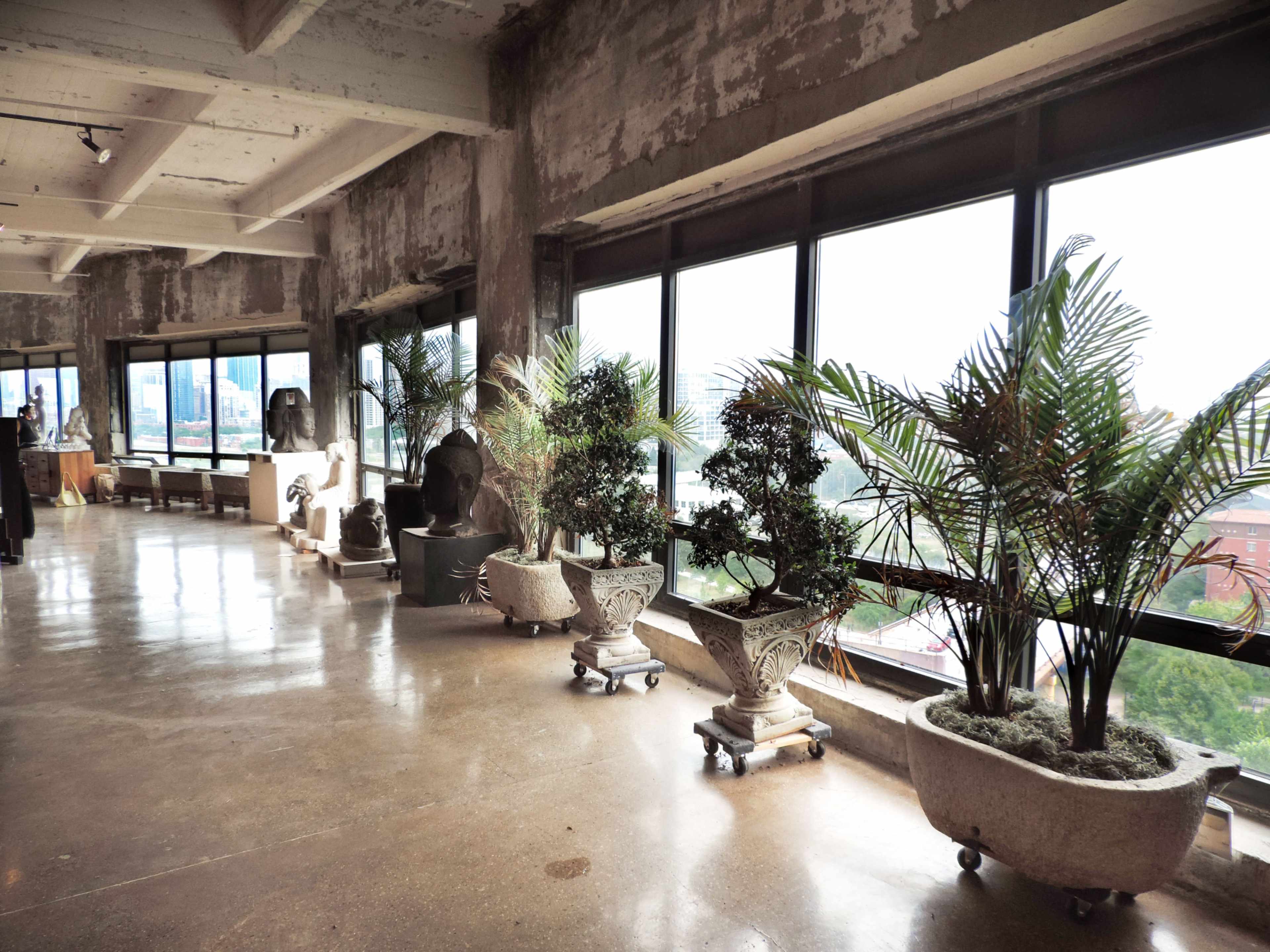 The image shows a spacious interior with large windows, potted plants, and sculptures along a polished concrete floor.