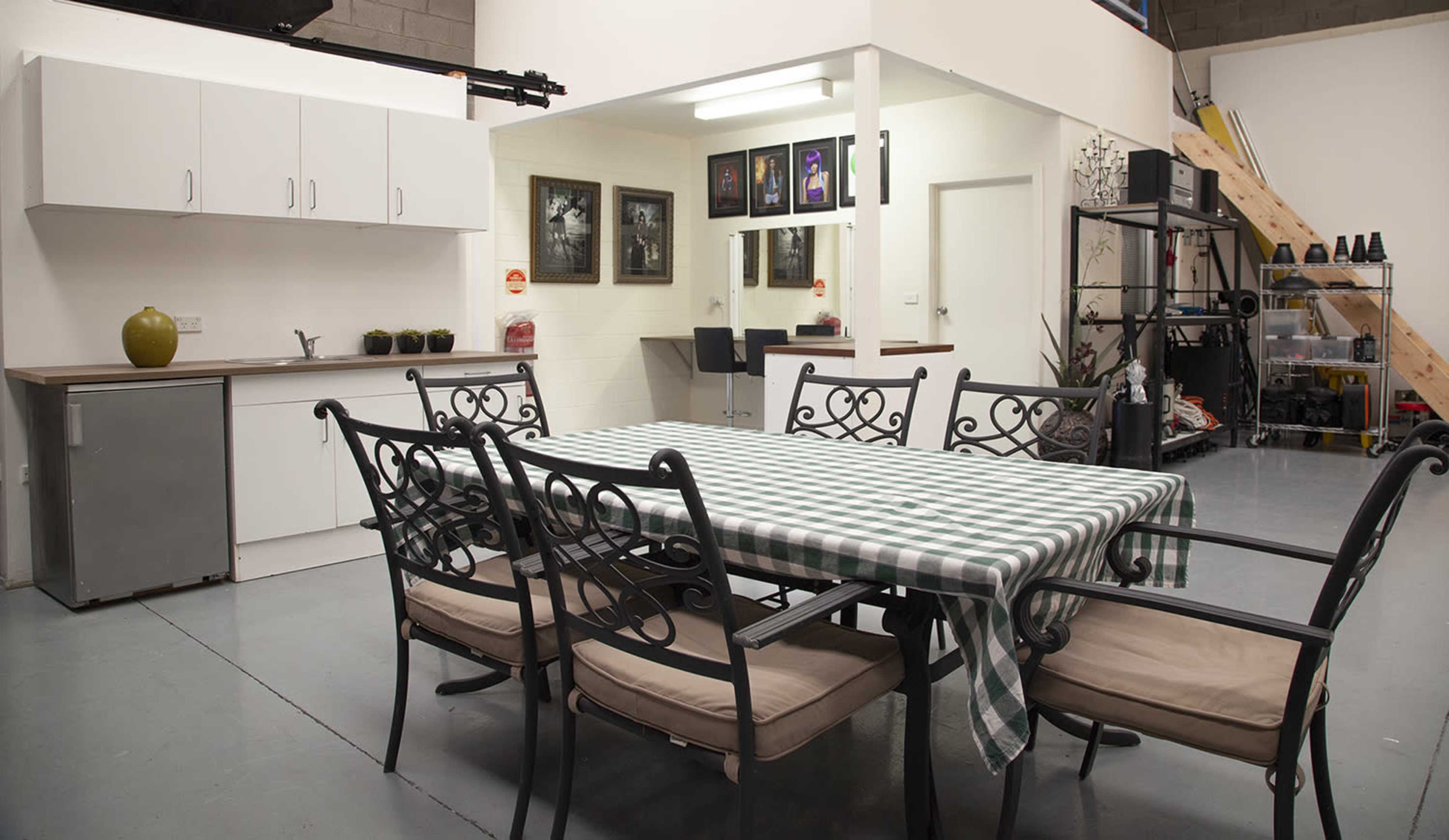A spacious interior featuring a dining table with a checkered tablecloth surrounded by metal chairs, a kitchenette with cabinets, and a staircase leading to an upper level.