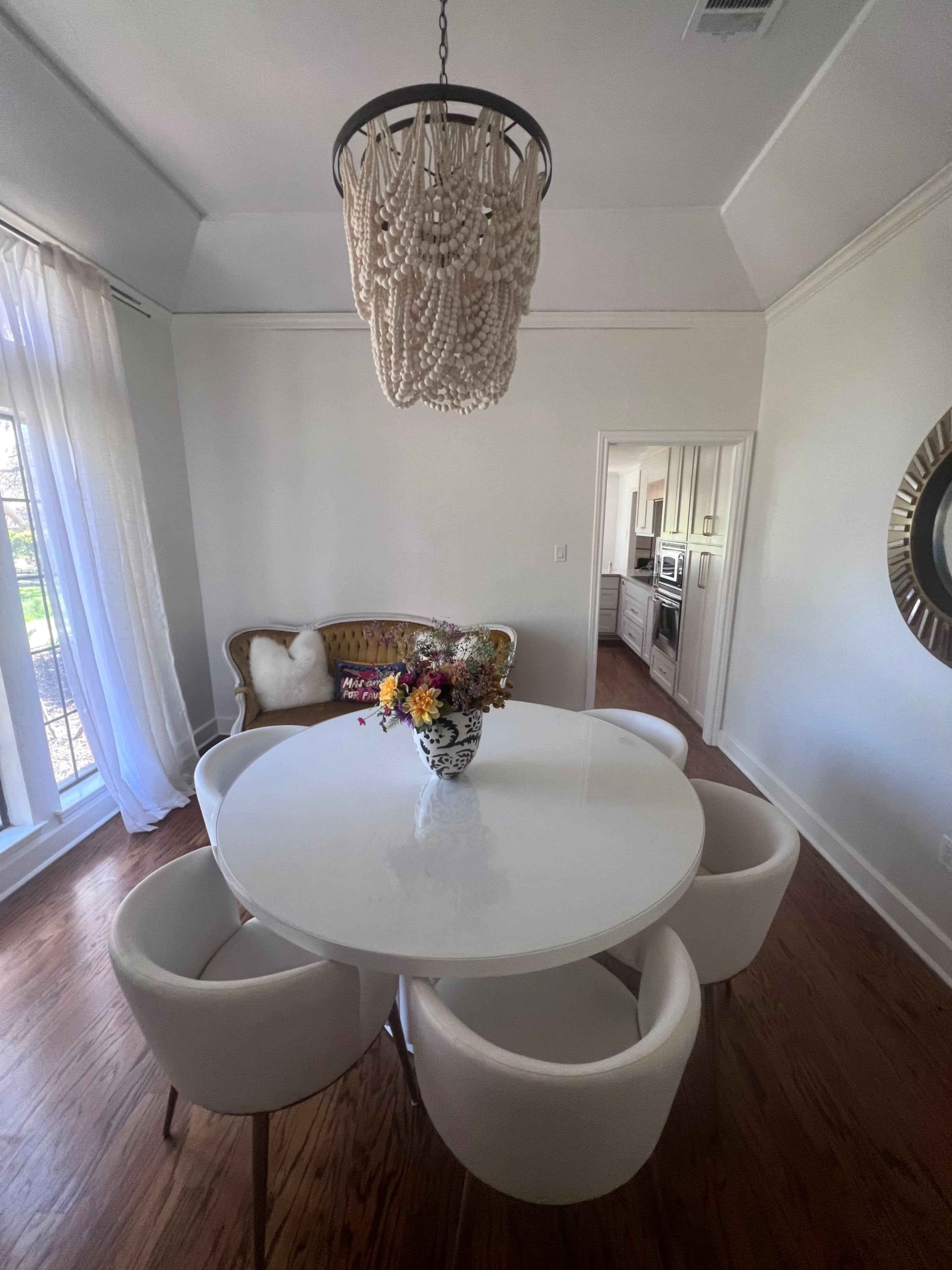 A round white dining table surrounded by four white chairs is placed in a well-lit room with a chandelier and a wicker sofa.