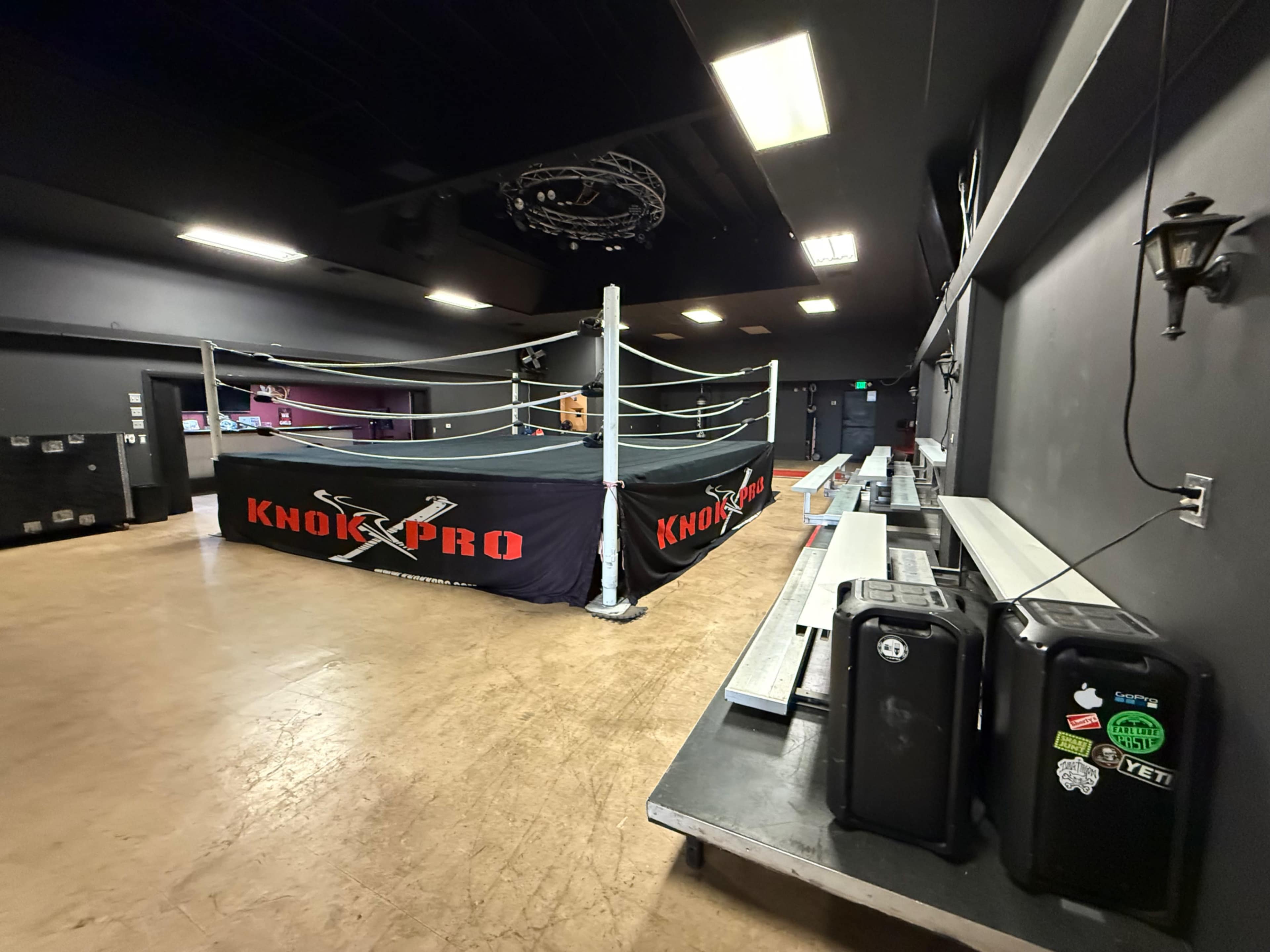 The image shows a wrestling ring set up in an indoor venue, surrounded by empty seating areas and equipment.