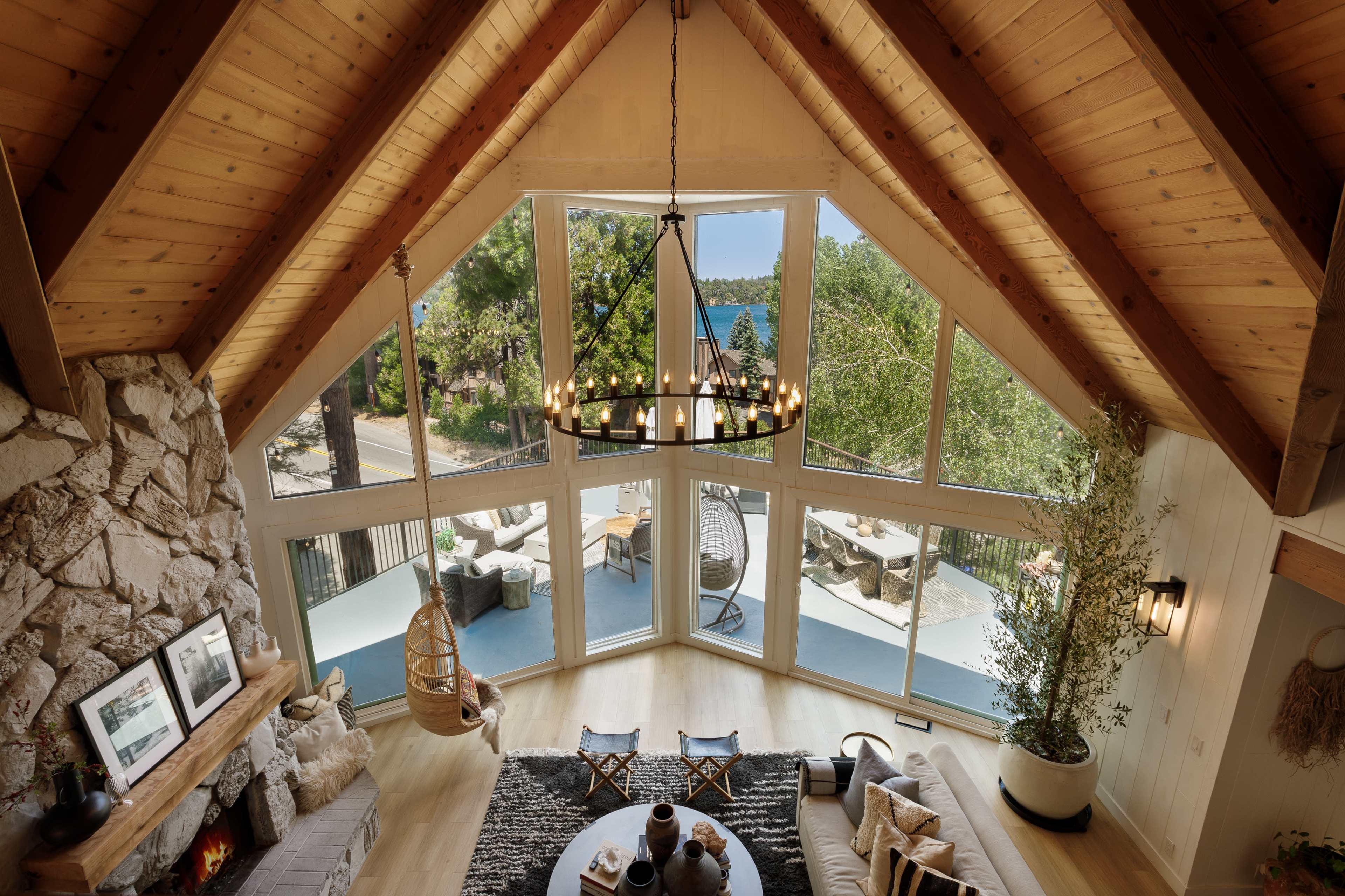 A spacious, modern living room with large windows offering a view of the outdoors, featuring a stone fireplace and a chandelier.