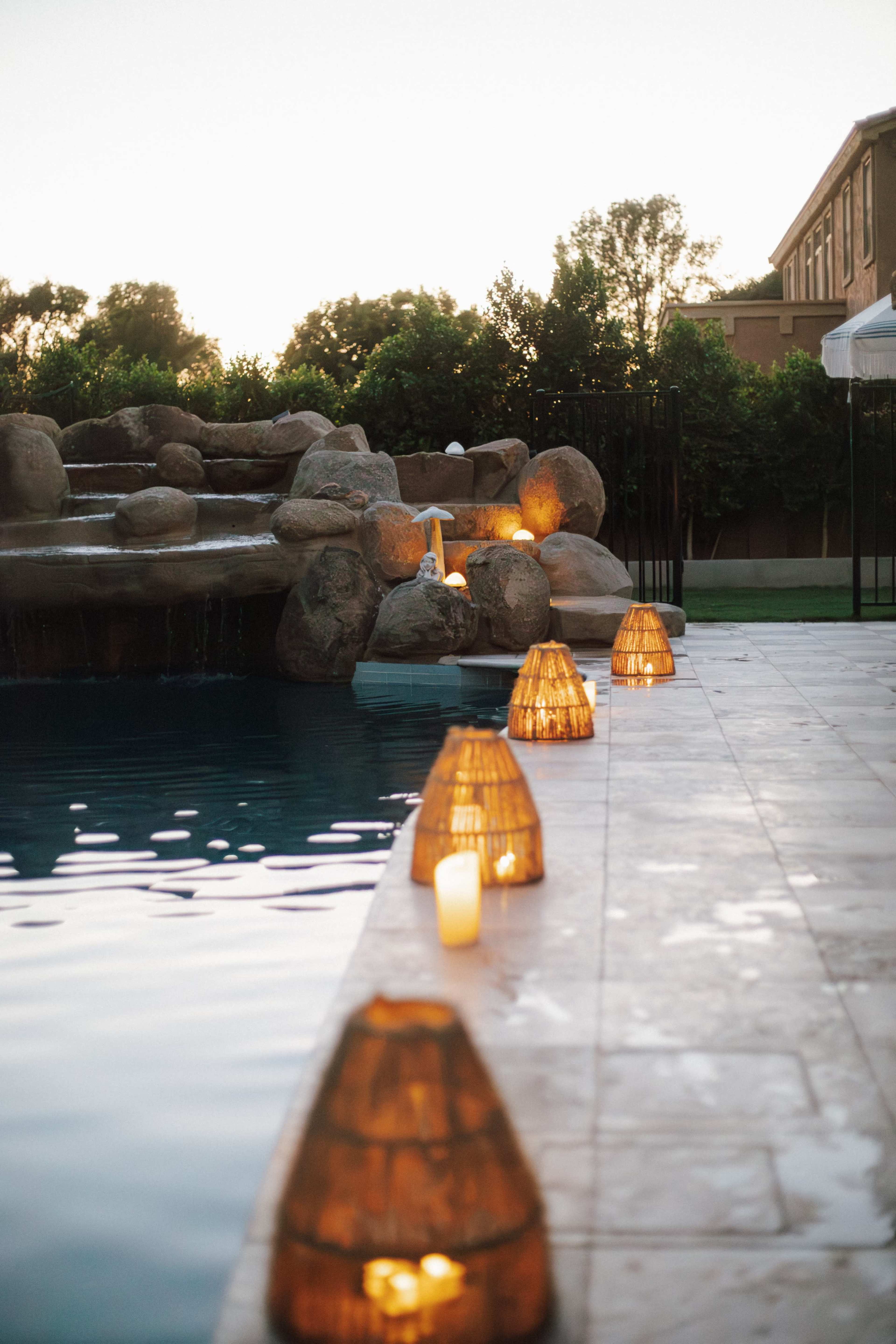 The image shows a swimming pool with a waterfall feature, lined with illuminated lanterns and surrounded by greenery and a building in the background.