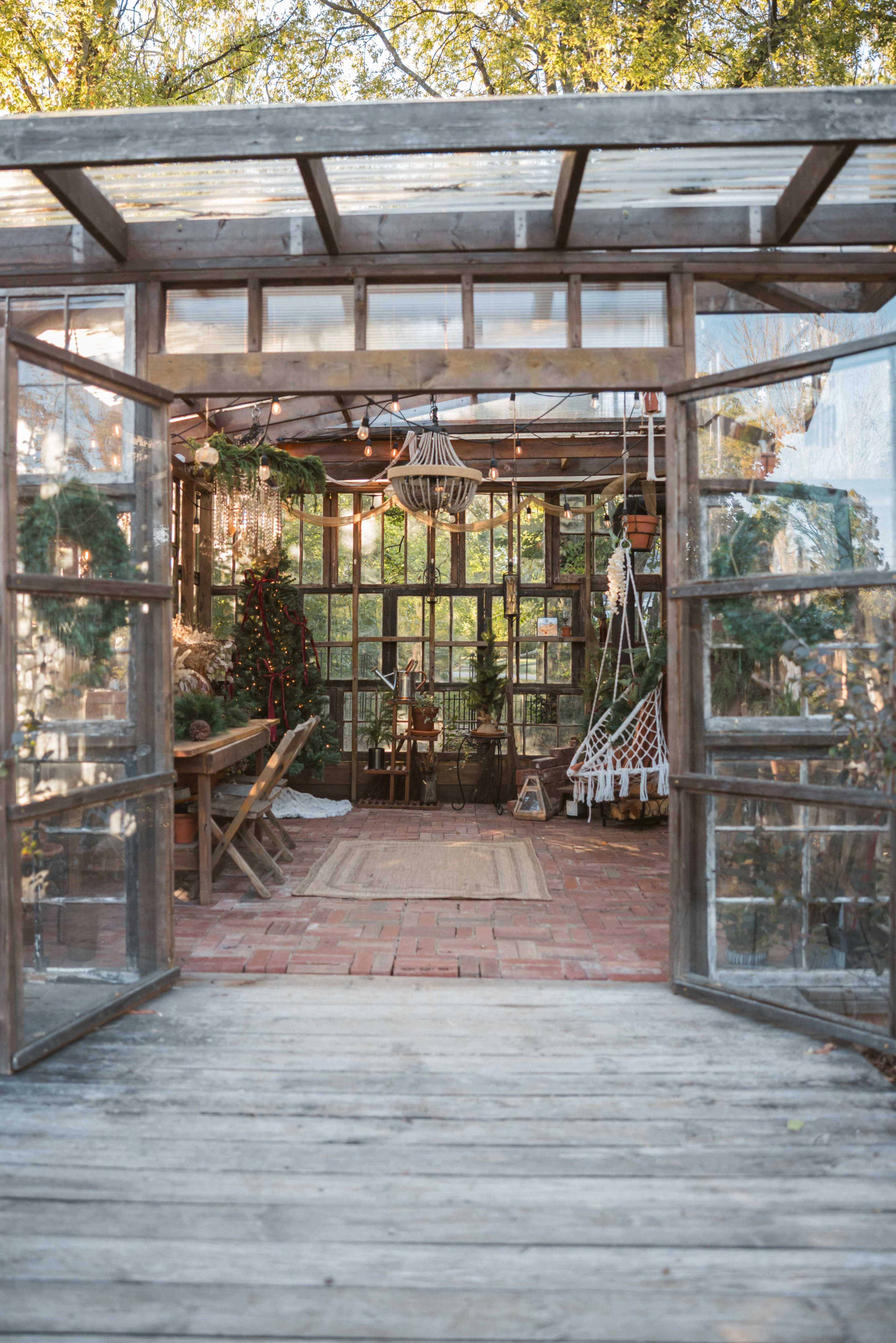 The image shows a wooden greenhouse with large glass doors open to reveal a cozy interior decorated with plants, lights, and rustic furniture.