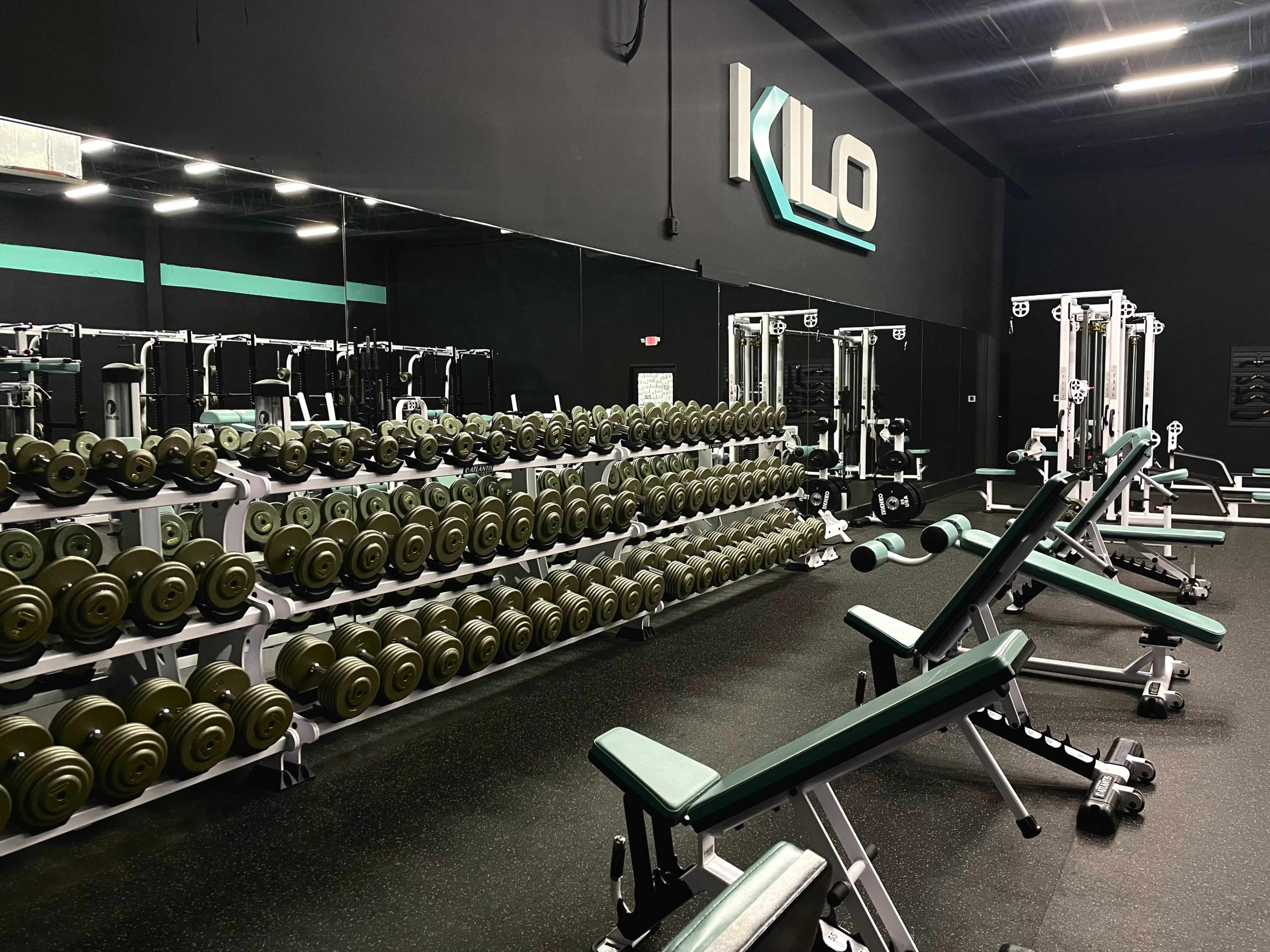 A well-organized gym featuring a long row of dumbbells, weight benches, and various strength training equipment against a black backdrop.