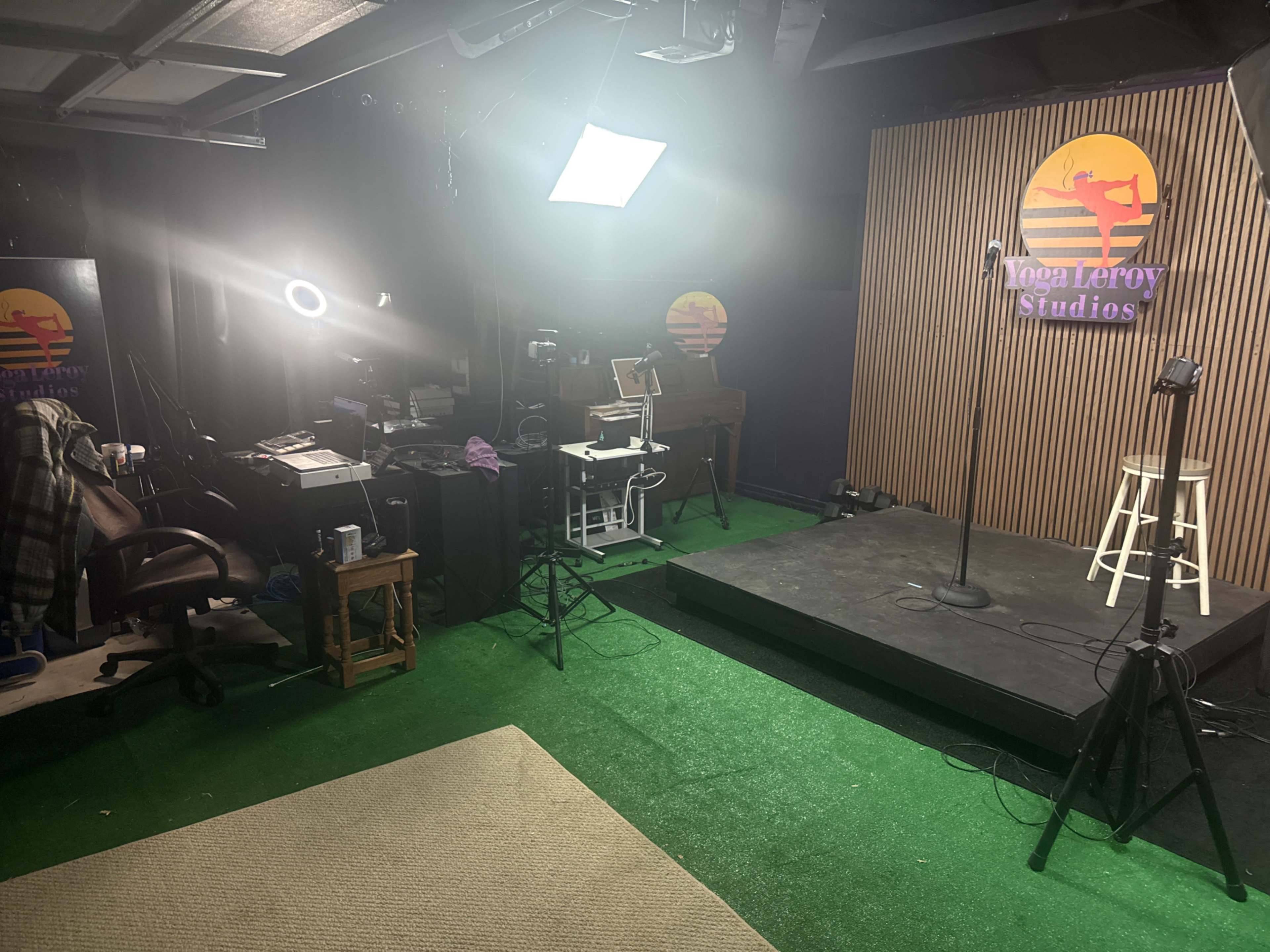 The image shows a recording studio setup with lights, a microphone stand, and various equipment arranged on a stage with artificial grass flooring.