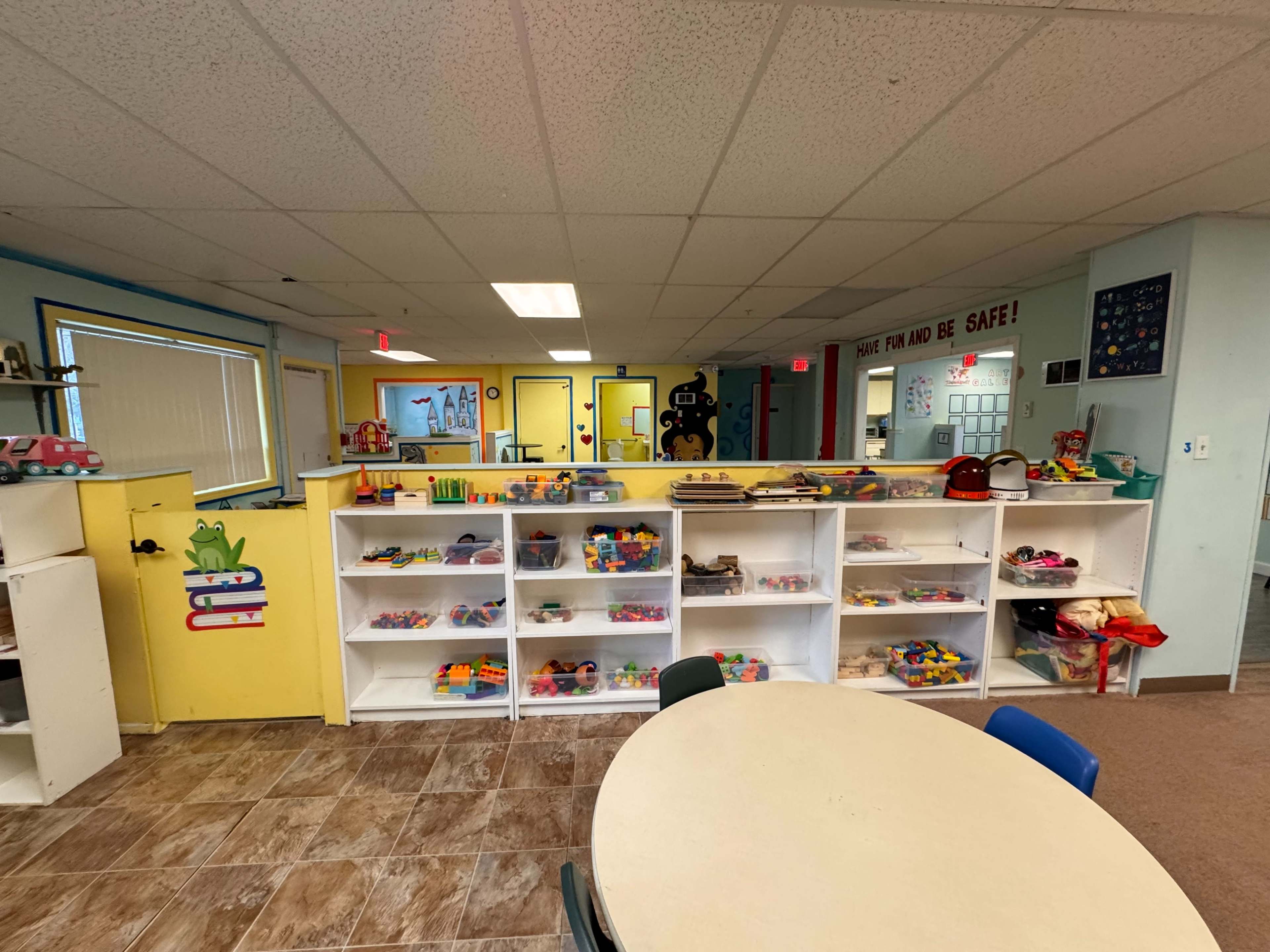 The image shows a brightly colored classroom with shelves filled with toys and a round table in the foreground.