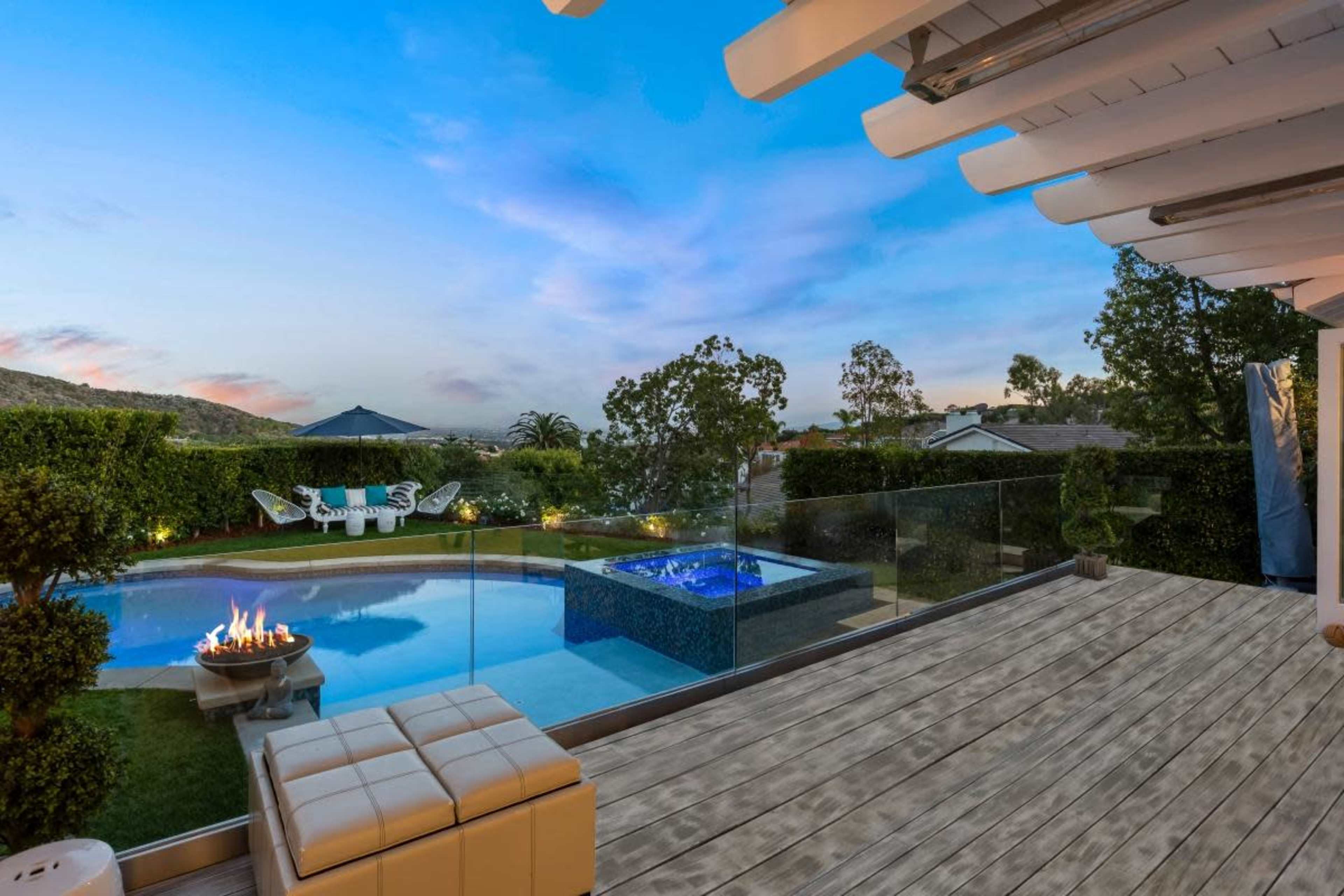 The image shows a patio with wooden flooring overlooking a swimming pool and a spa, surrounded by greenery and trees under a blue sky.