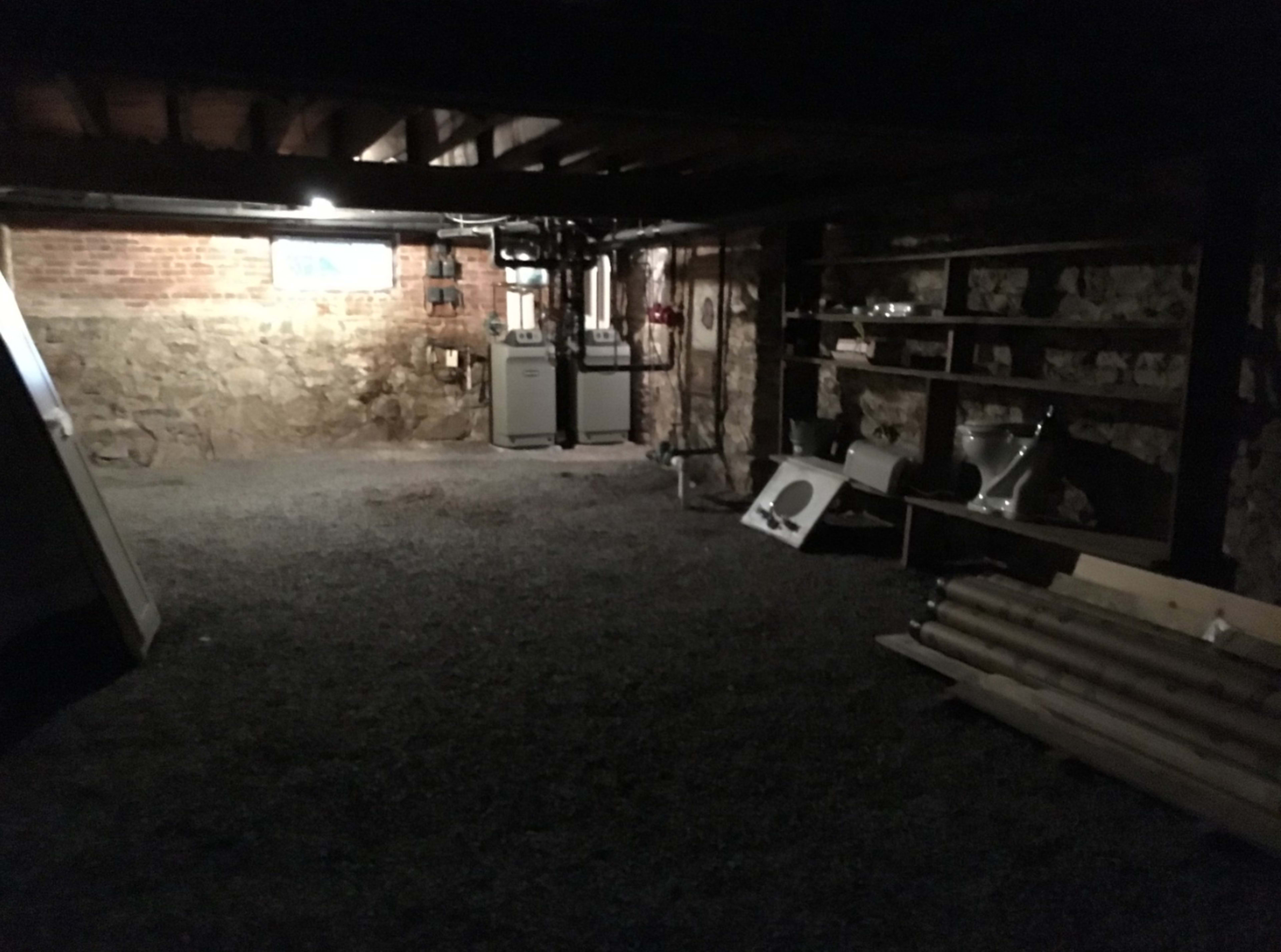 A dimly lit basement with gravel flooring, exposed stone walls, a few appliances, and shelves containing various items.