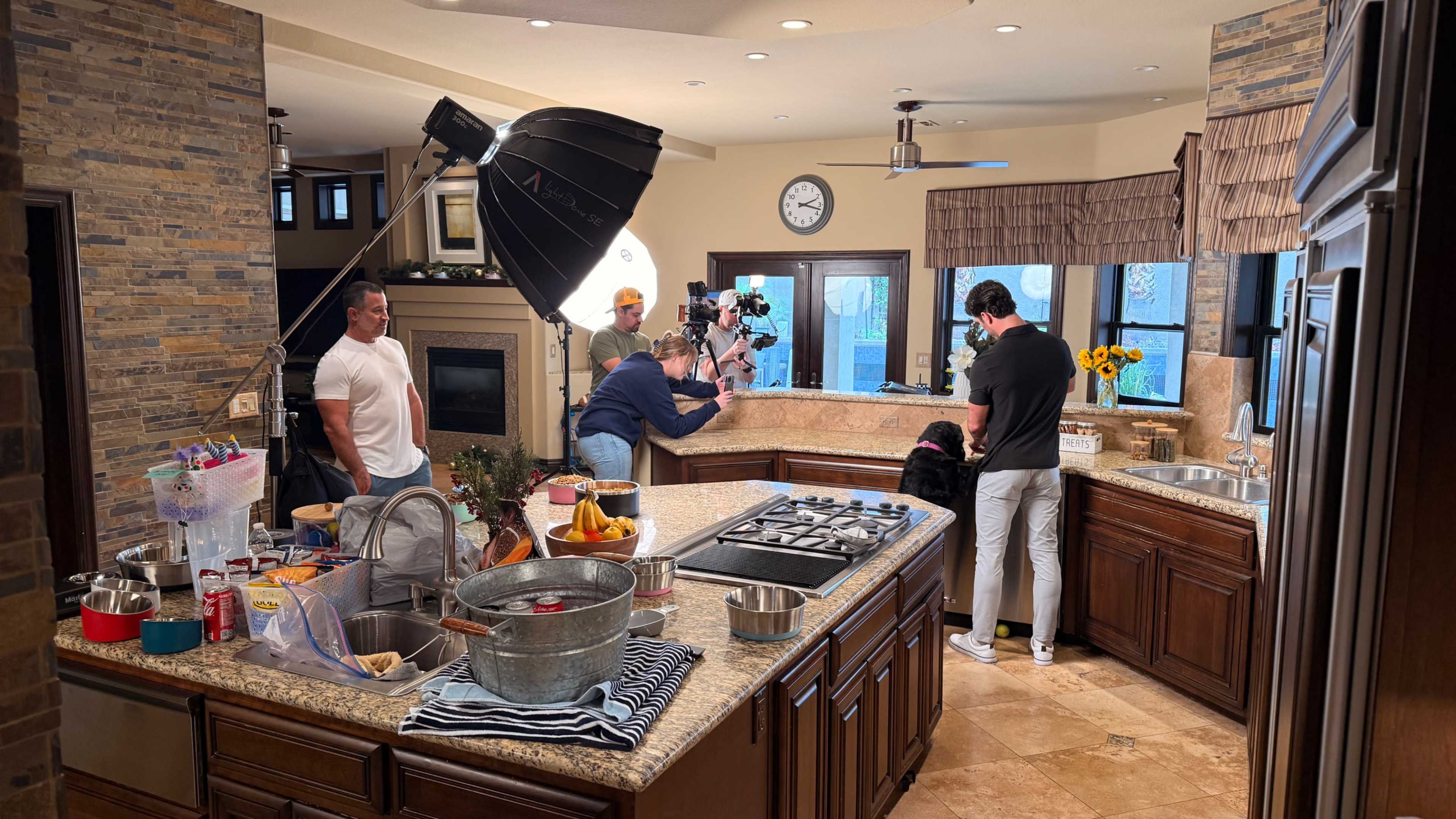 A film crew sets up equipment in a spacious kitchen where two individuals are engaged in an activity, surrounded by various kitchen tools and ingredients.