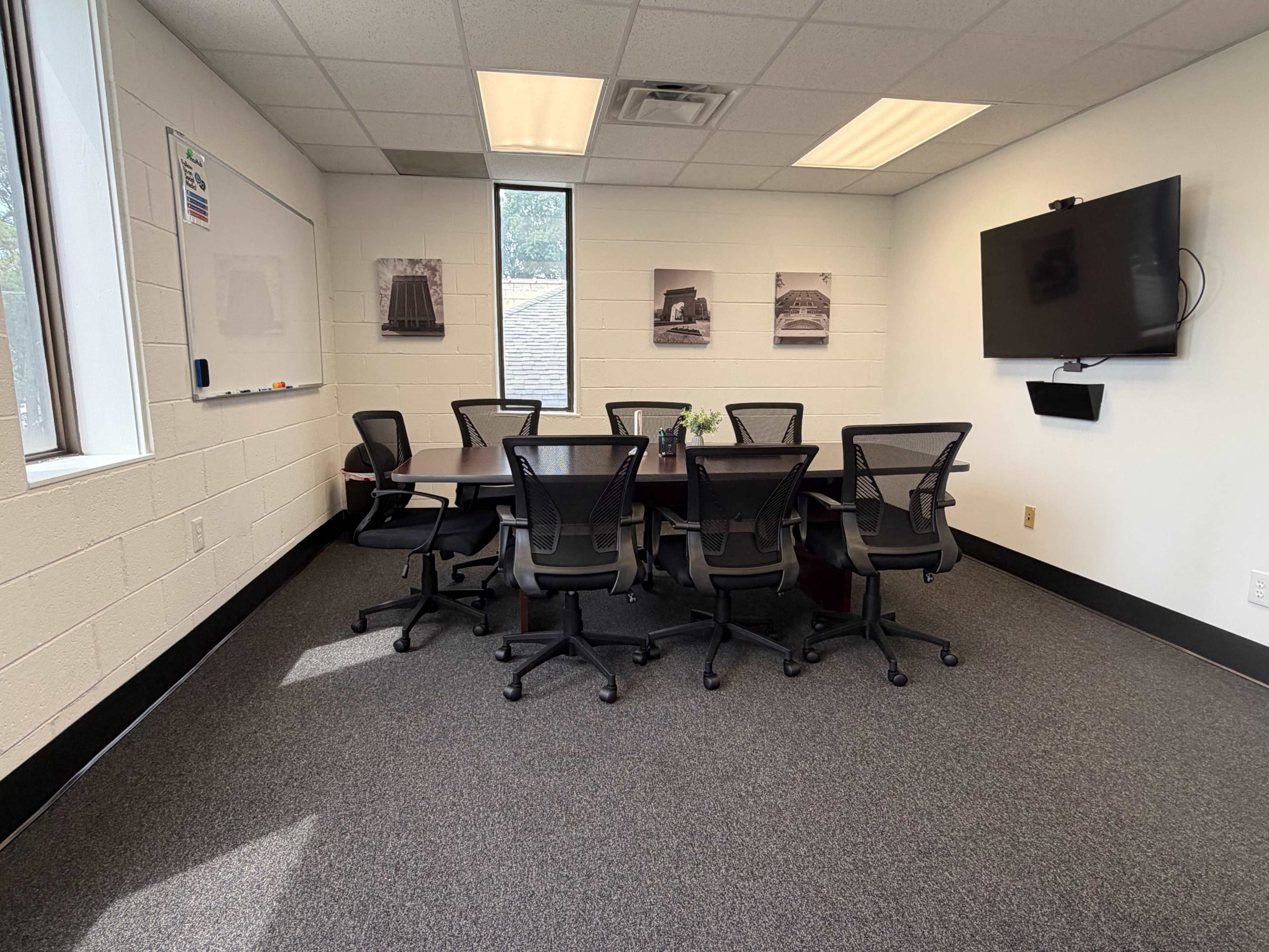 The image shows a meeting room with a large table surrounded by eight ergonomic chairs, a wall-mounted TV, and several black-and-white photographs on the walls.