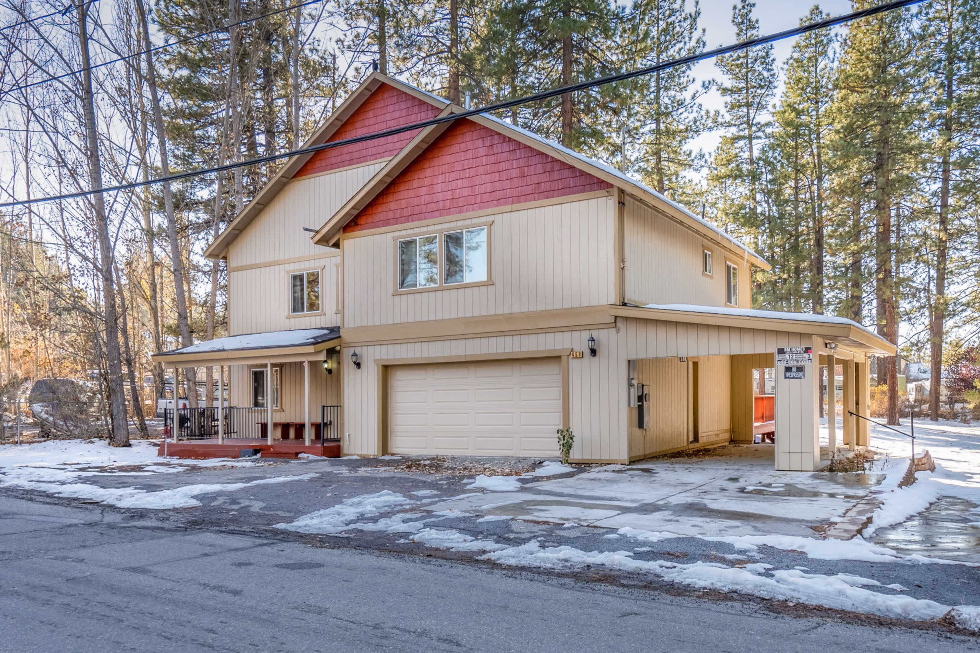 A two-story house with a light-colored exterior and red accents, situated on a snowy street lined with tall pine trees.