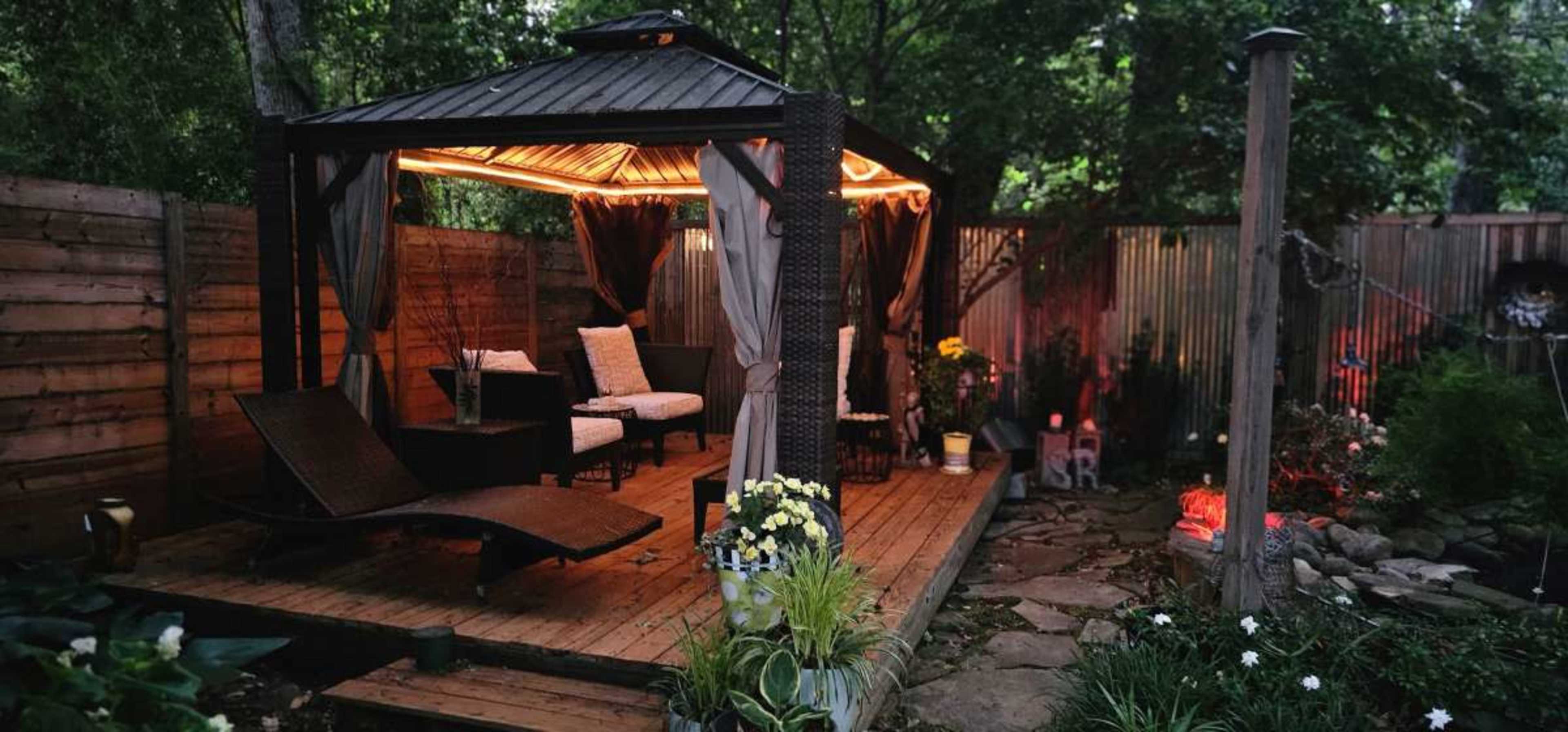 The image shows a garden gazebo with seating and decorative lighting surrounded by greenery and stone pathways.