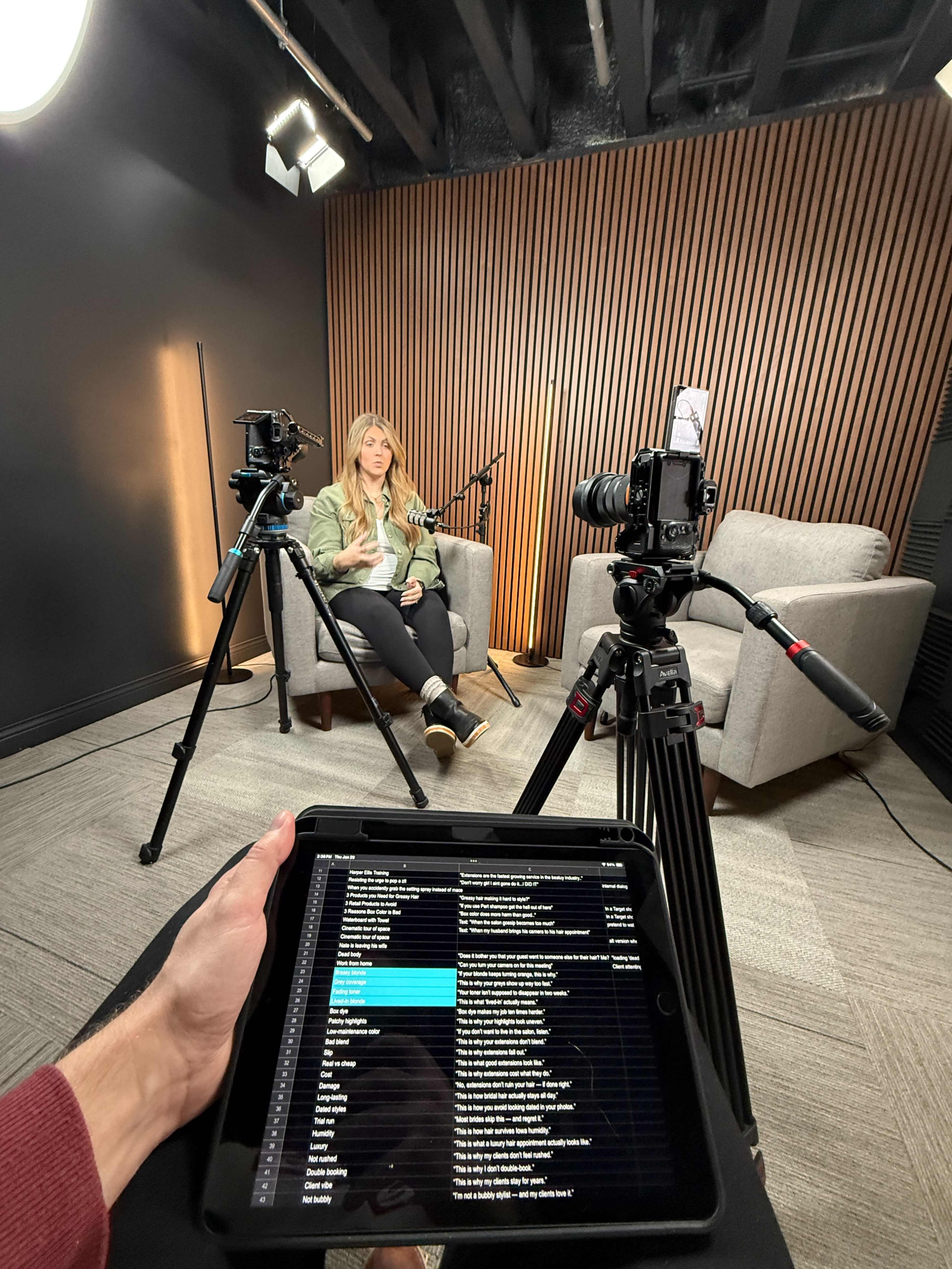 A person sits on a gray couch in a studio, while two cameras on tripods and a person holding an iPad with notes are visible in the foreground.