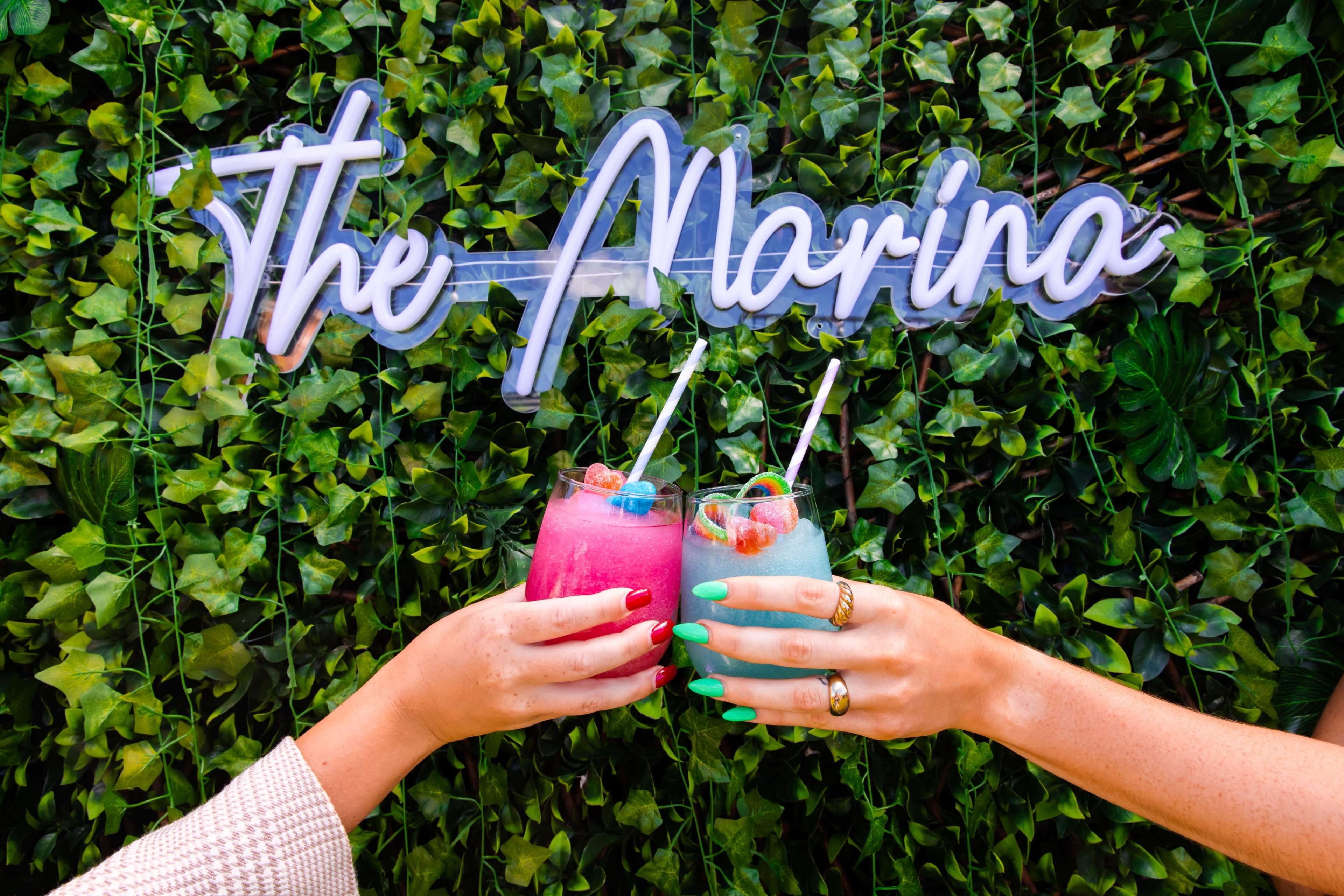 Two hands clink colorful frozen drinks in front of a greenery-covered backdrop with the text "The Alarina."
