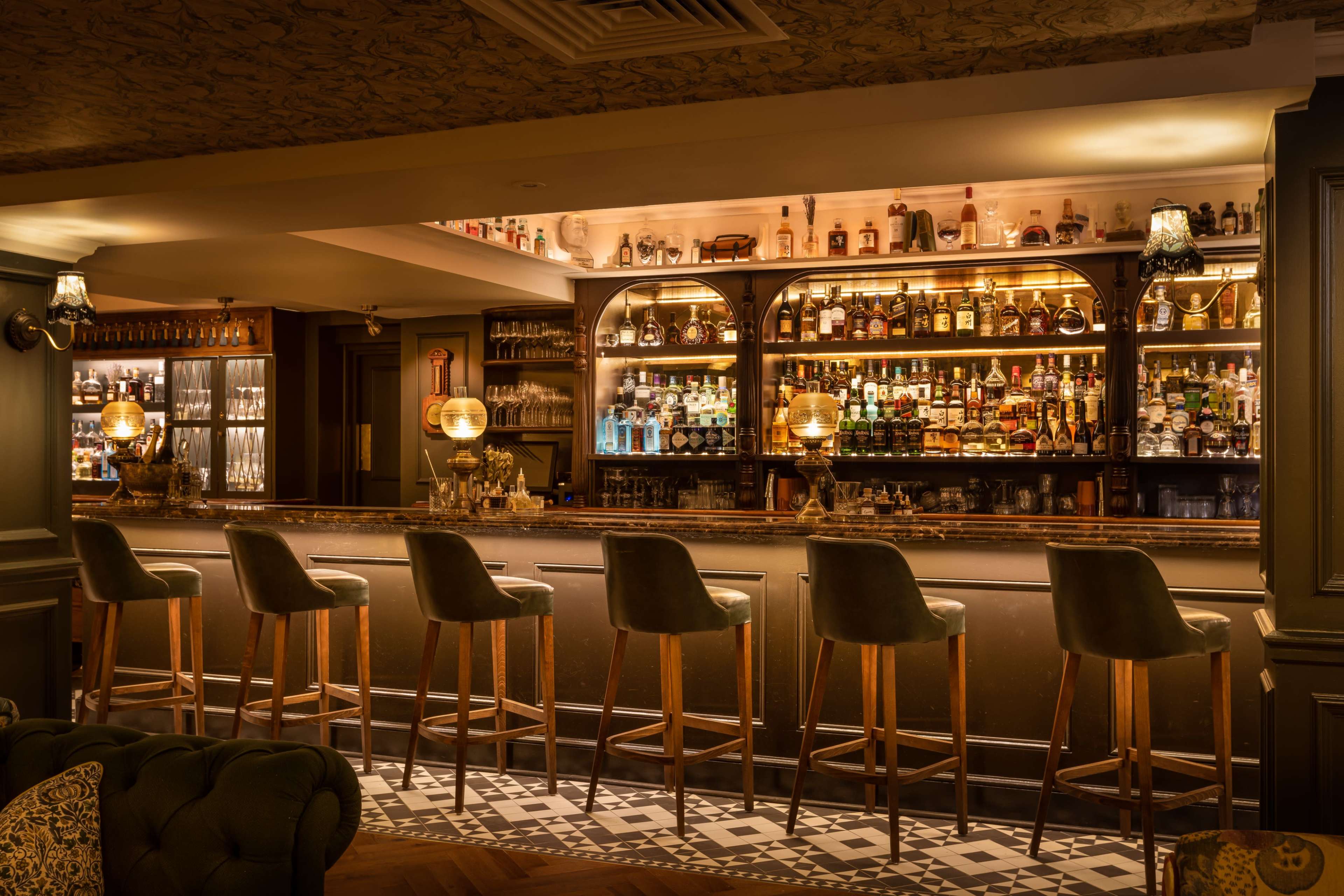 A dimly lit bar with a polished counter, high stools, and shelves lined with various bottles of liquor.
