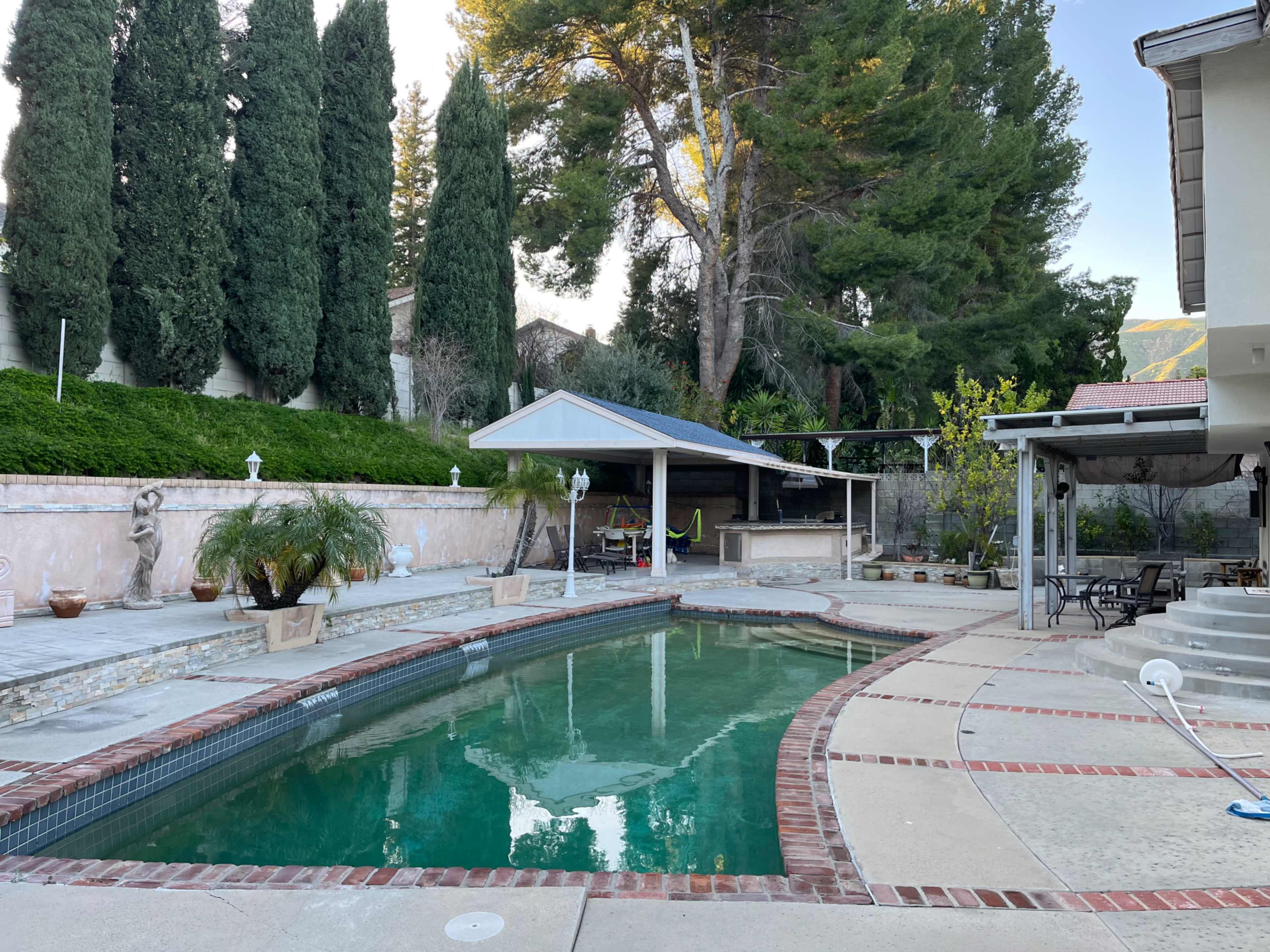 A backyard features a swimming pool surrounded by concrete and stone pathways, with tall trees and a covered patio in the background.
