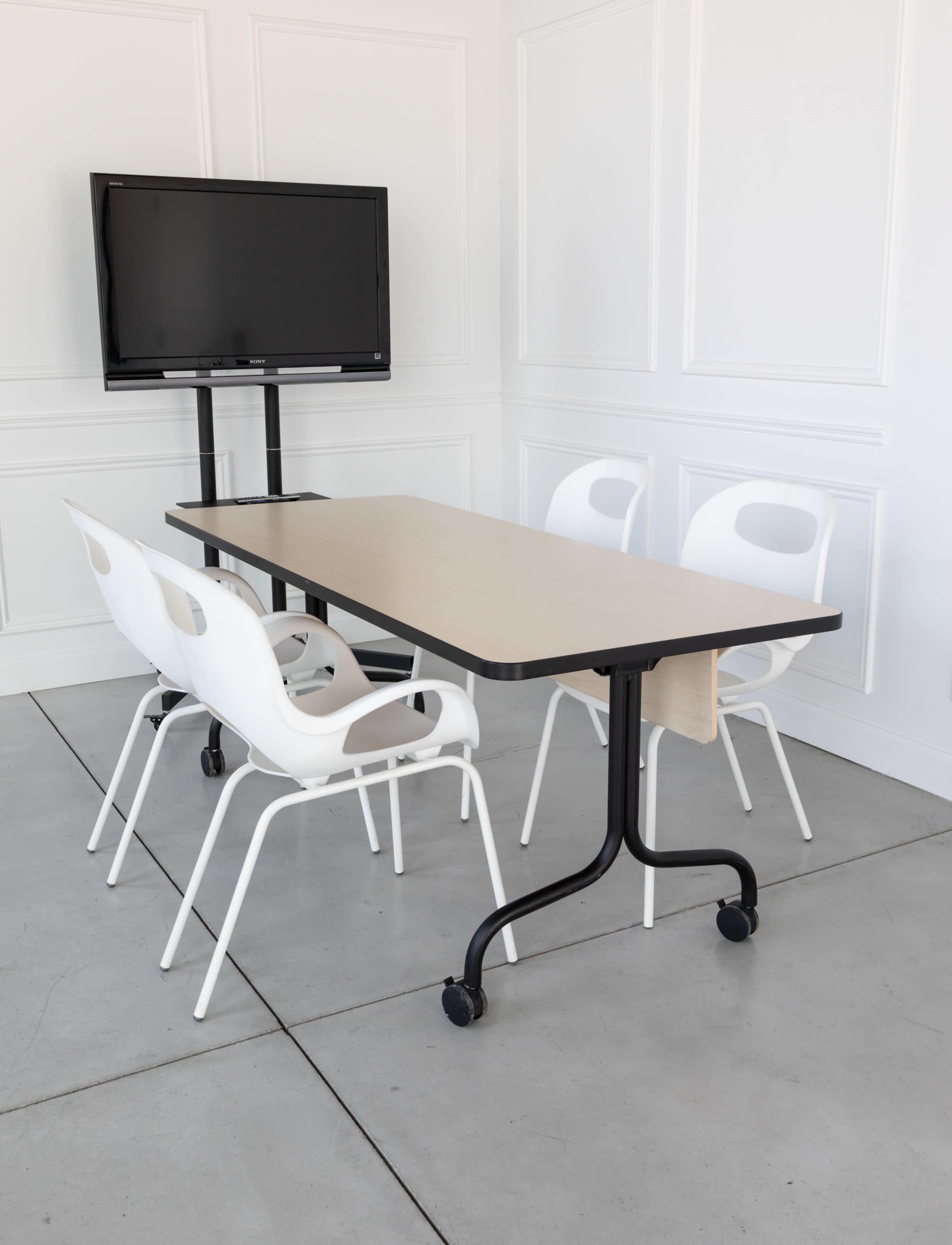 A modern conference room setup features a light wooden table with a black frame and four white chairs, alongside a mounted TV on a stand.