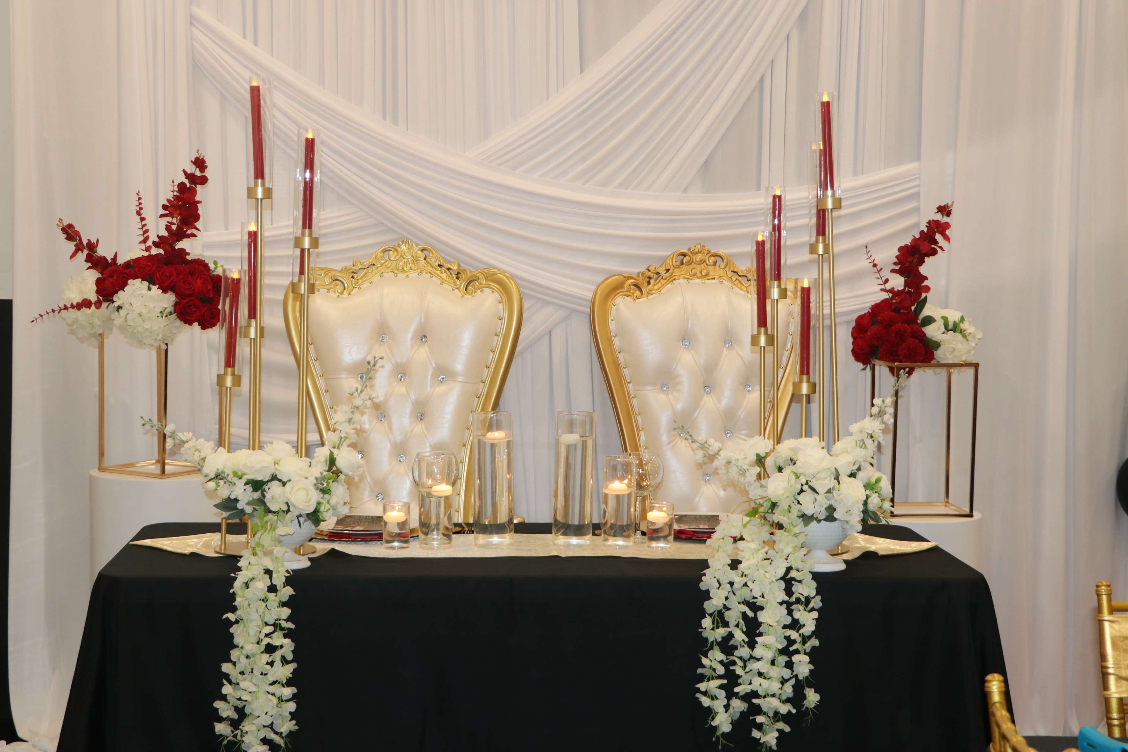 The image shows an elegantly decorated table with two ornate white and gold chairs, flanked by tall candle holders and floral arrangements featuring red roses and white flowers.