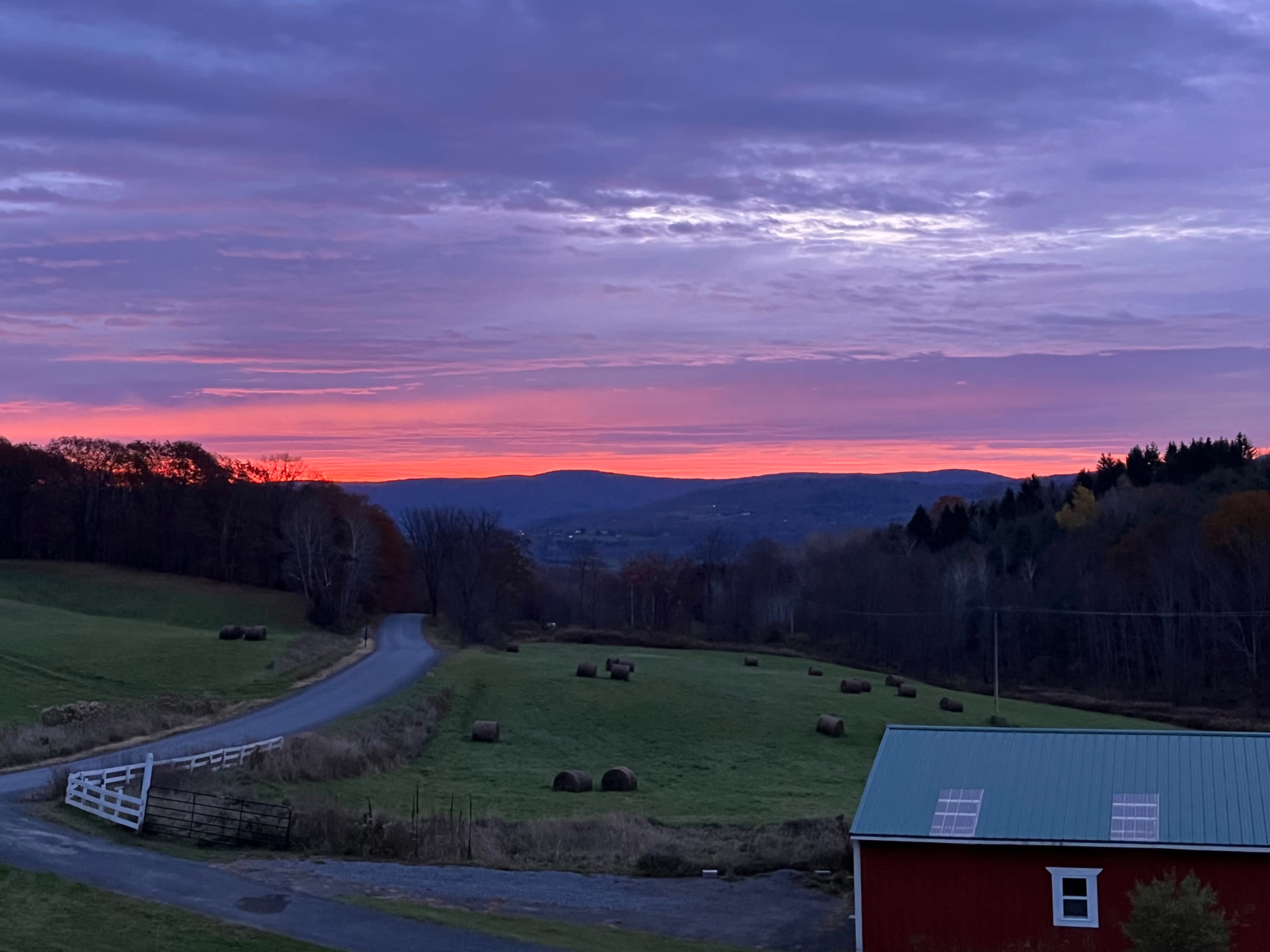 The scene shows a rural landscape with rolled hay bales scattered in a green field, a winding road, and a red barn under a colorful sunset sky.