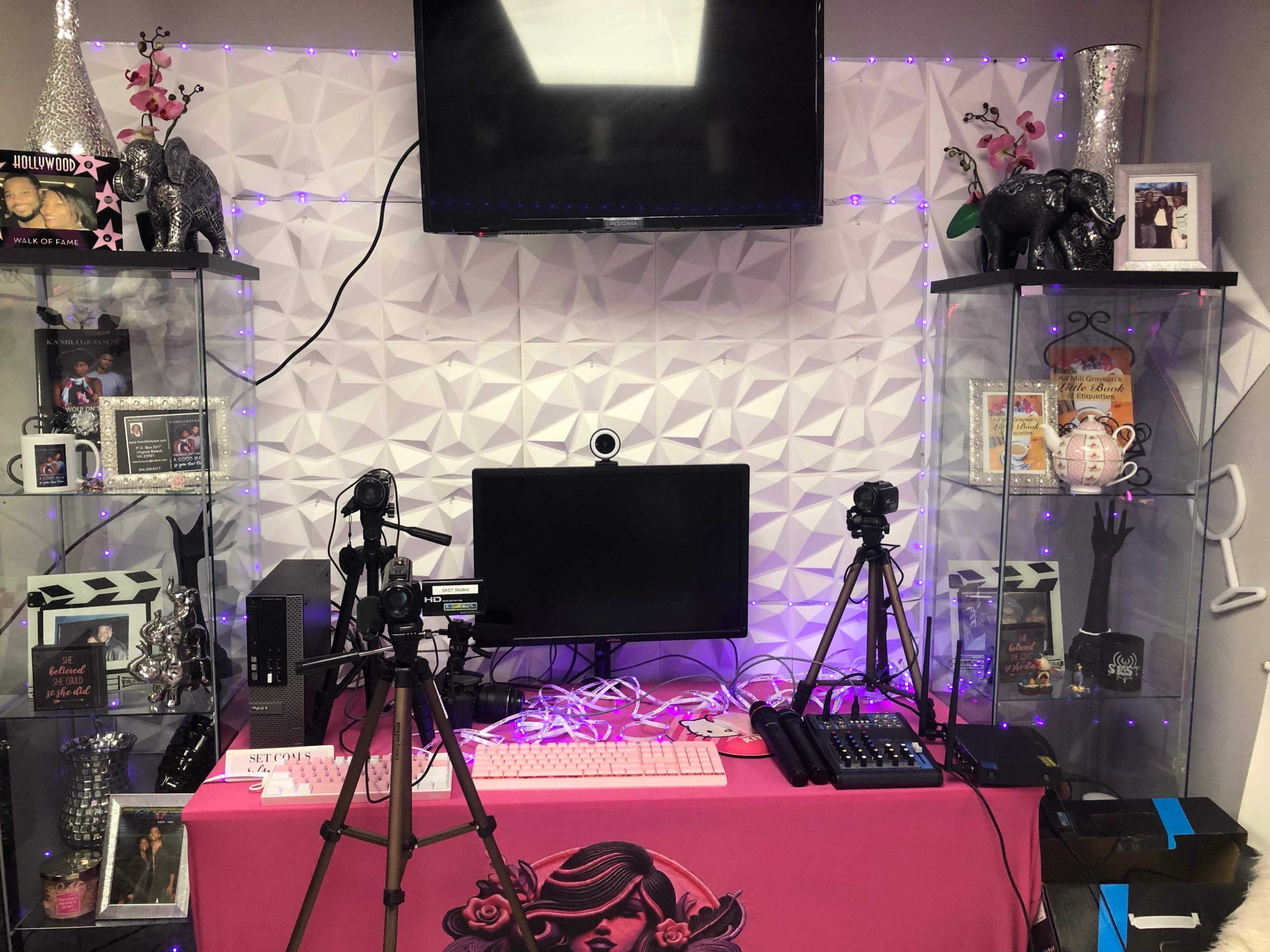 The image shows a setup for video recording with a computer monitor, multiple cameras on tripods, and decorative lighting against a patterned backdrop.