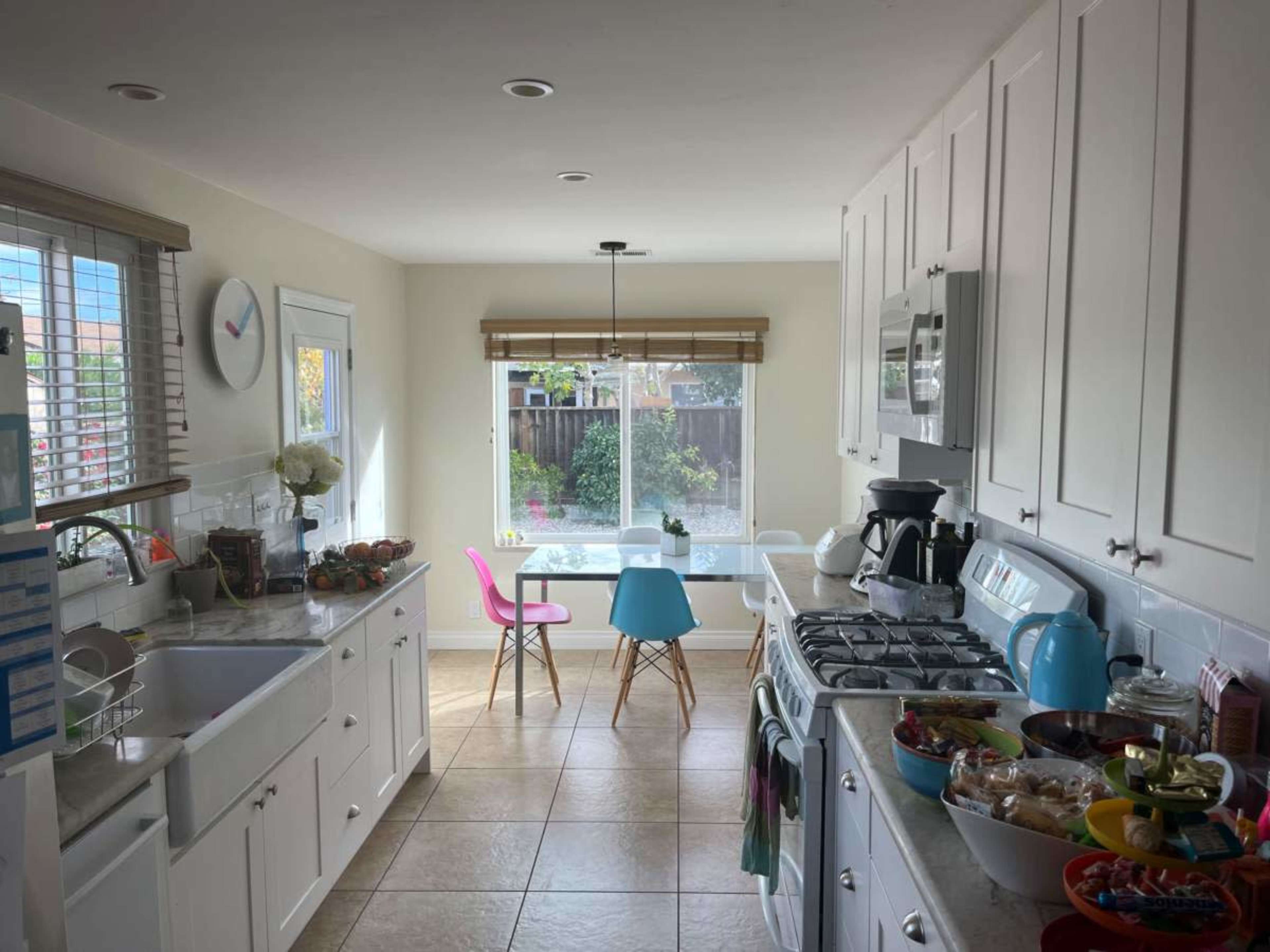 A bright kitchen features white cabinets, a gas stove, a small table with colorful chairs, and a variety of food items laid out on the counter.