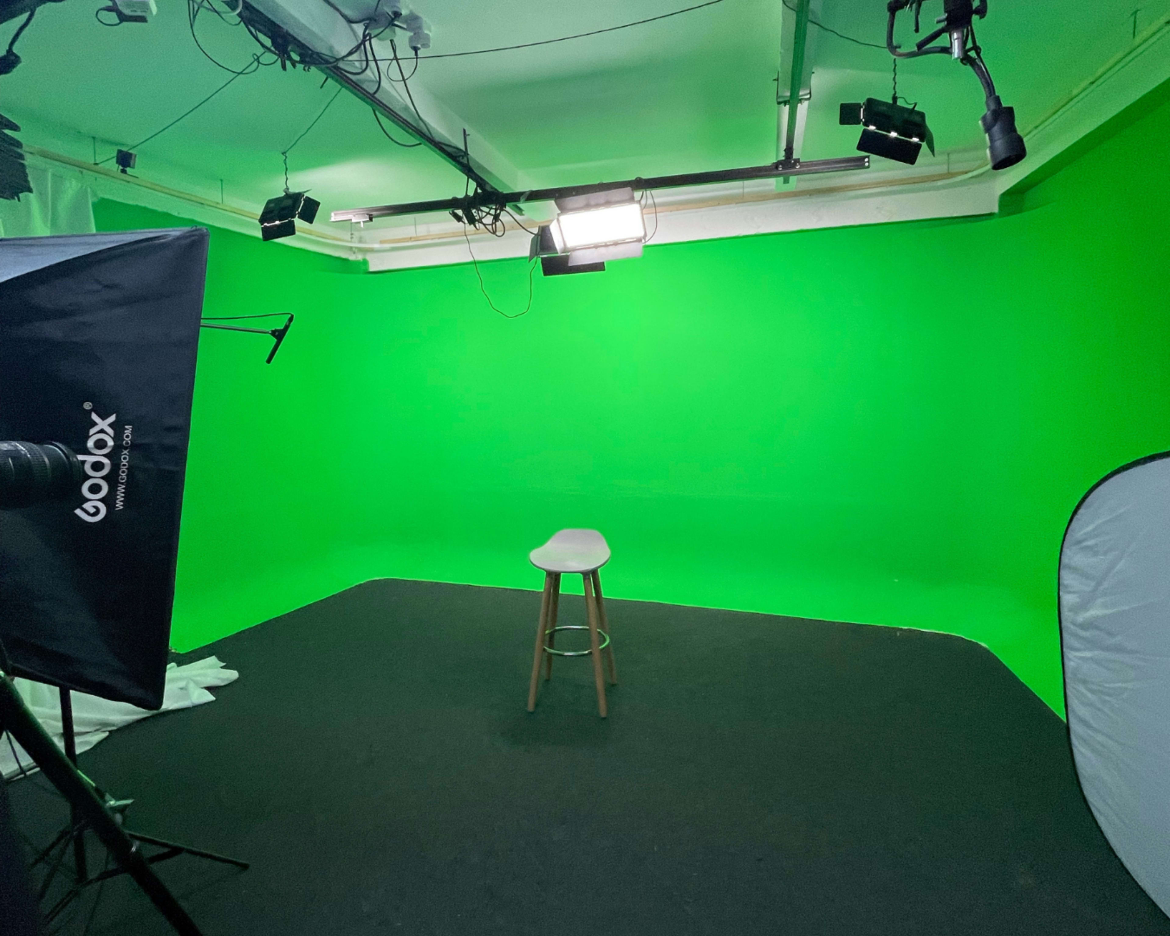 The image shows a green screen studio with a single wooden stool placed in the center and various lighting equipment surrounding it.