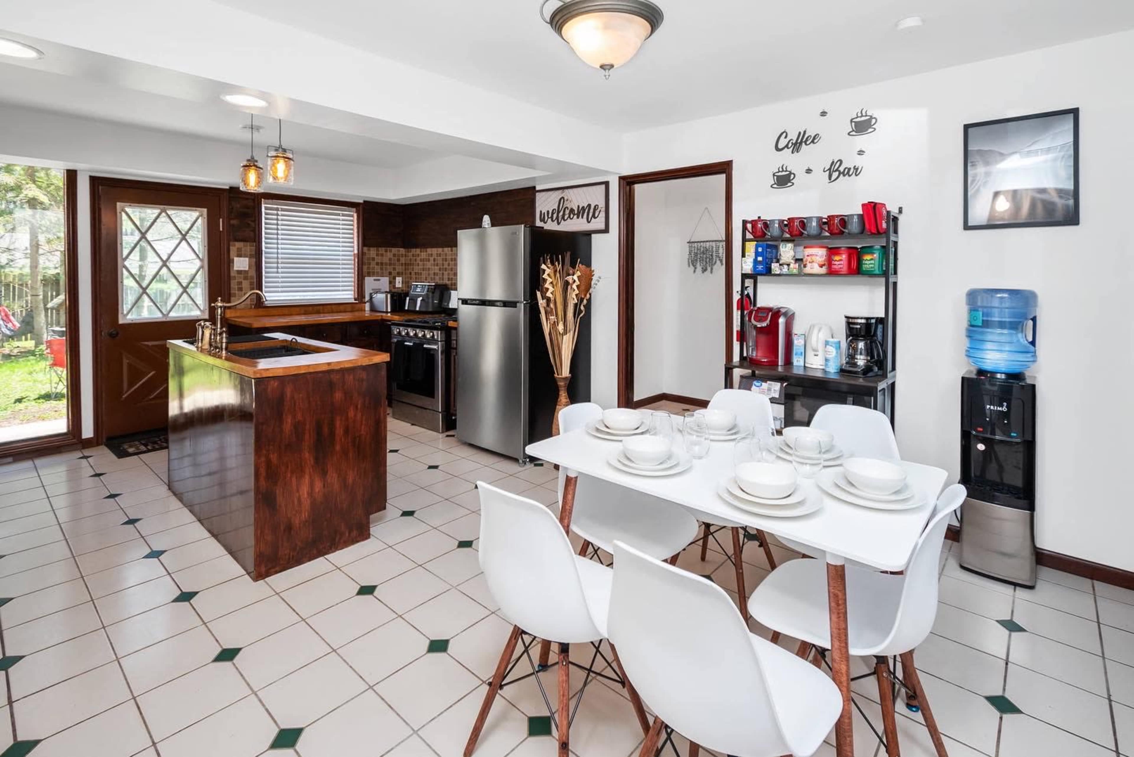 A modern kitchen with a dining table set for four, a stainless steel refrigerator, and a coffee bar along the wall.