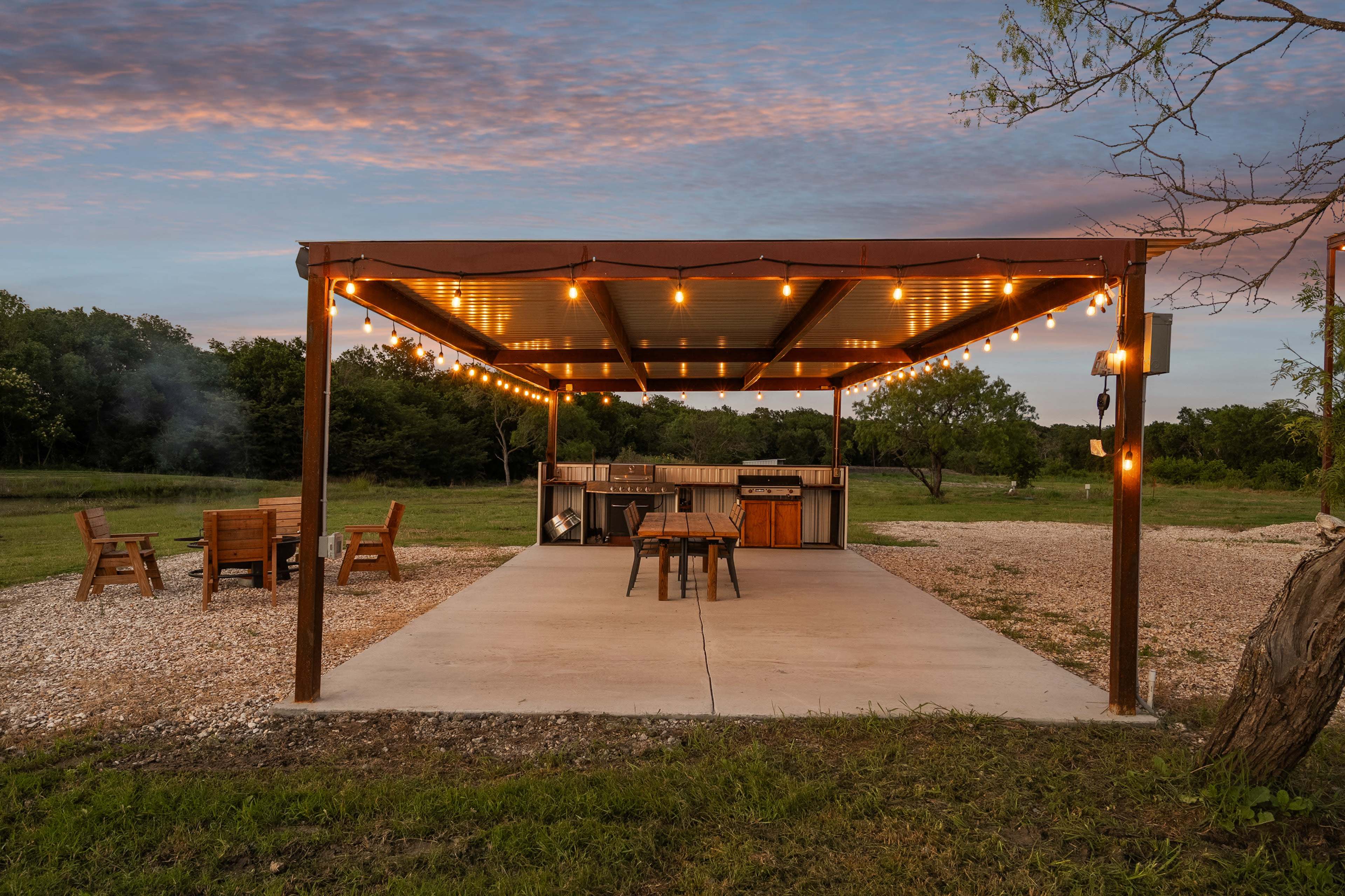 The image shows a covered outdoor patio with string lights, a barbecue grill, a dining table, and seating around a fire pit in a grassy area.