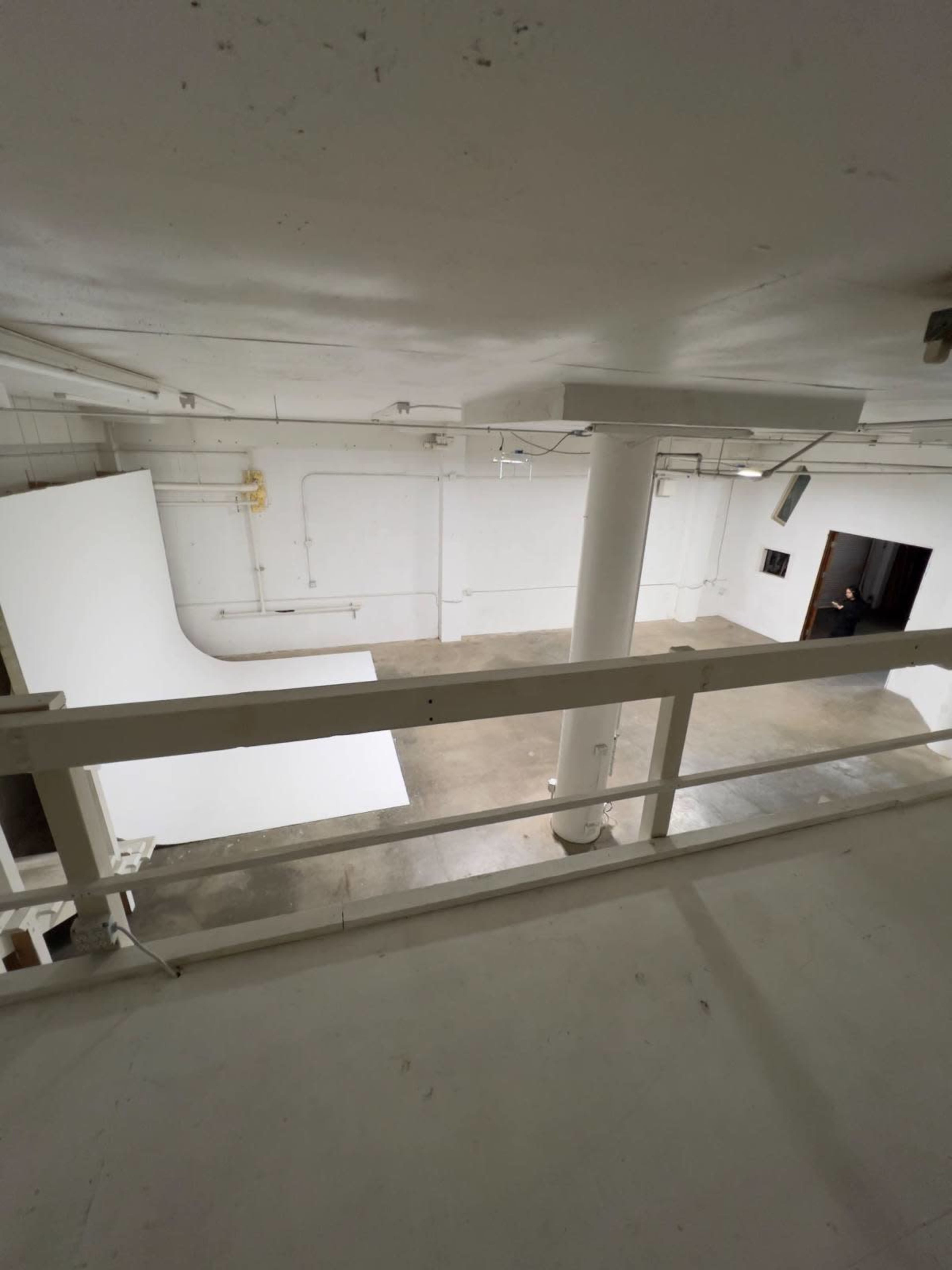 The image shows an empty warehouse space with a large white backdrop and a concrete floor, viewed from an upper level.