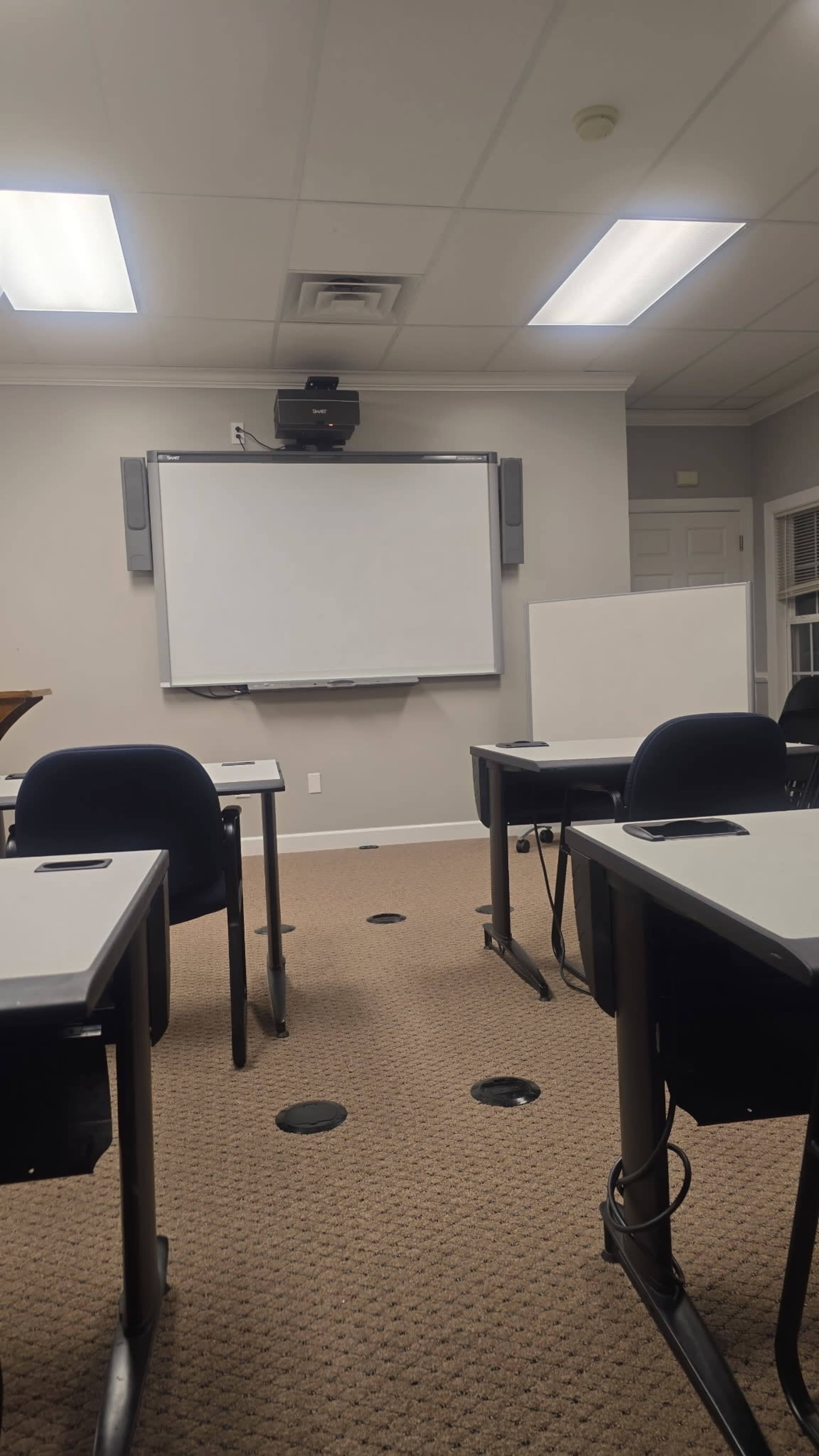 The image shows a classroom with multiple desks arranged in rows facing a blank whiteboard at the front of the room.