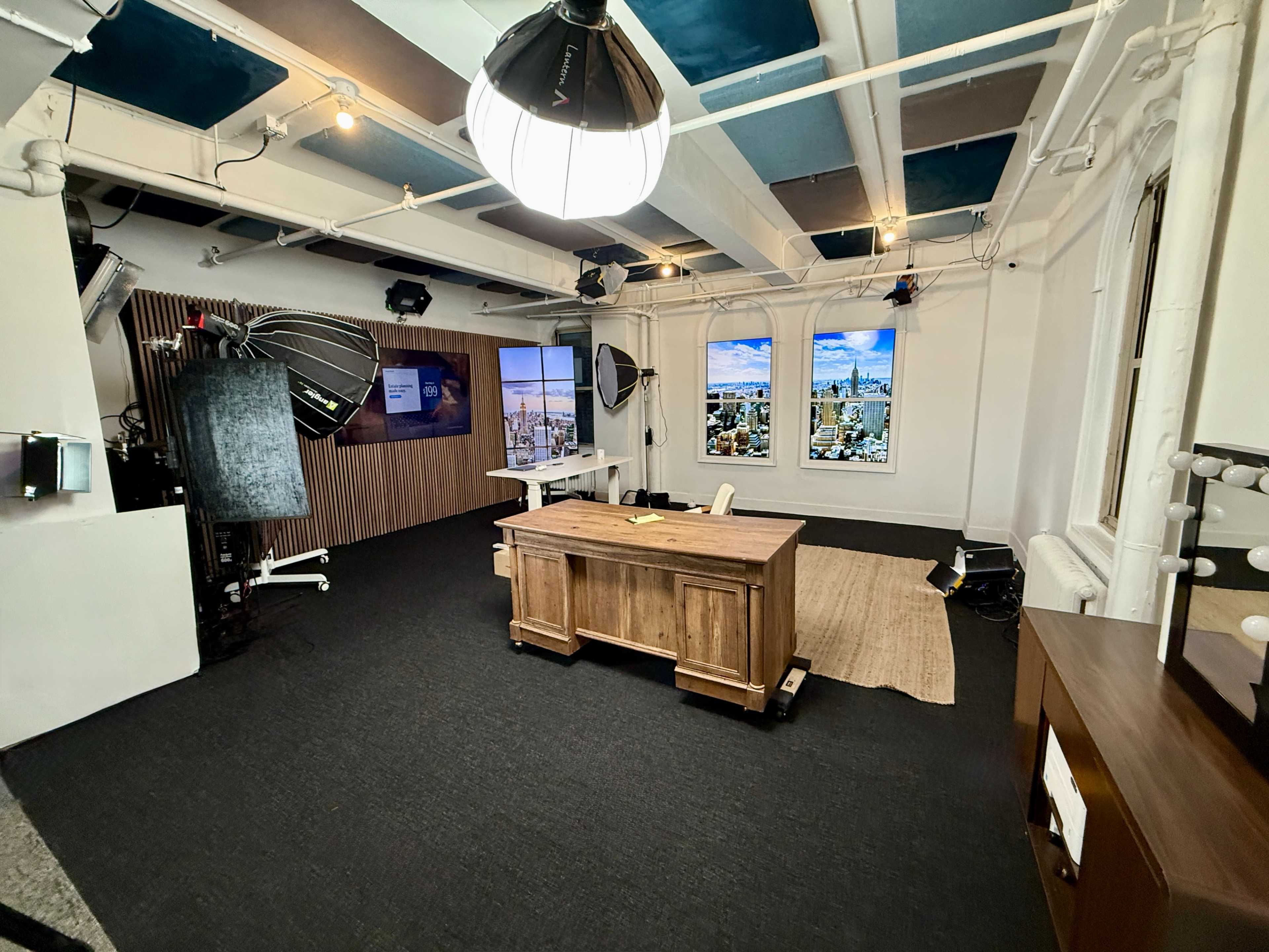 The image shows a well-lit studio space featuring a wooden desk, a large window with a city view, lighting equipment, and a textured rug on the floor.