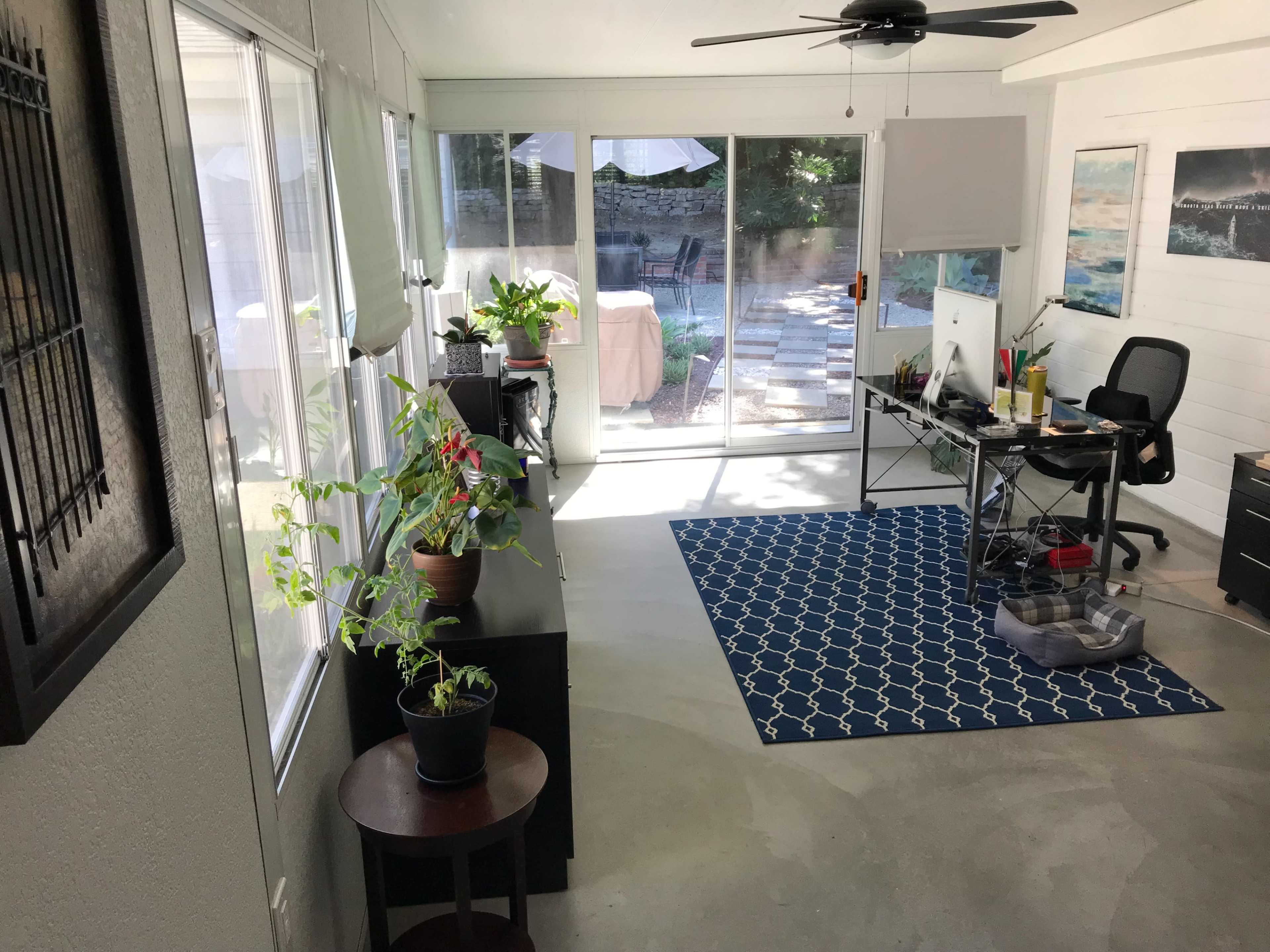 The image shows a bright, modern room with a desk, chair, and potted plants, overlooking a patio area through large windows.