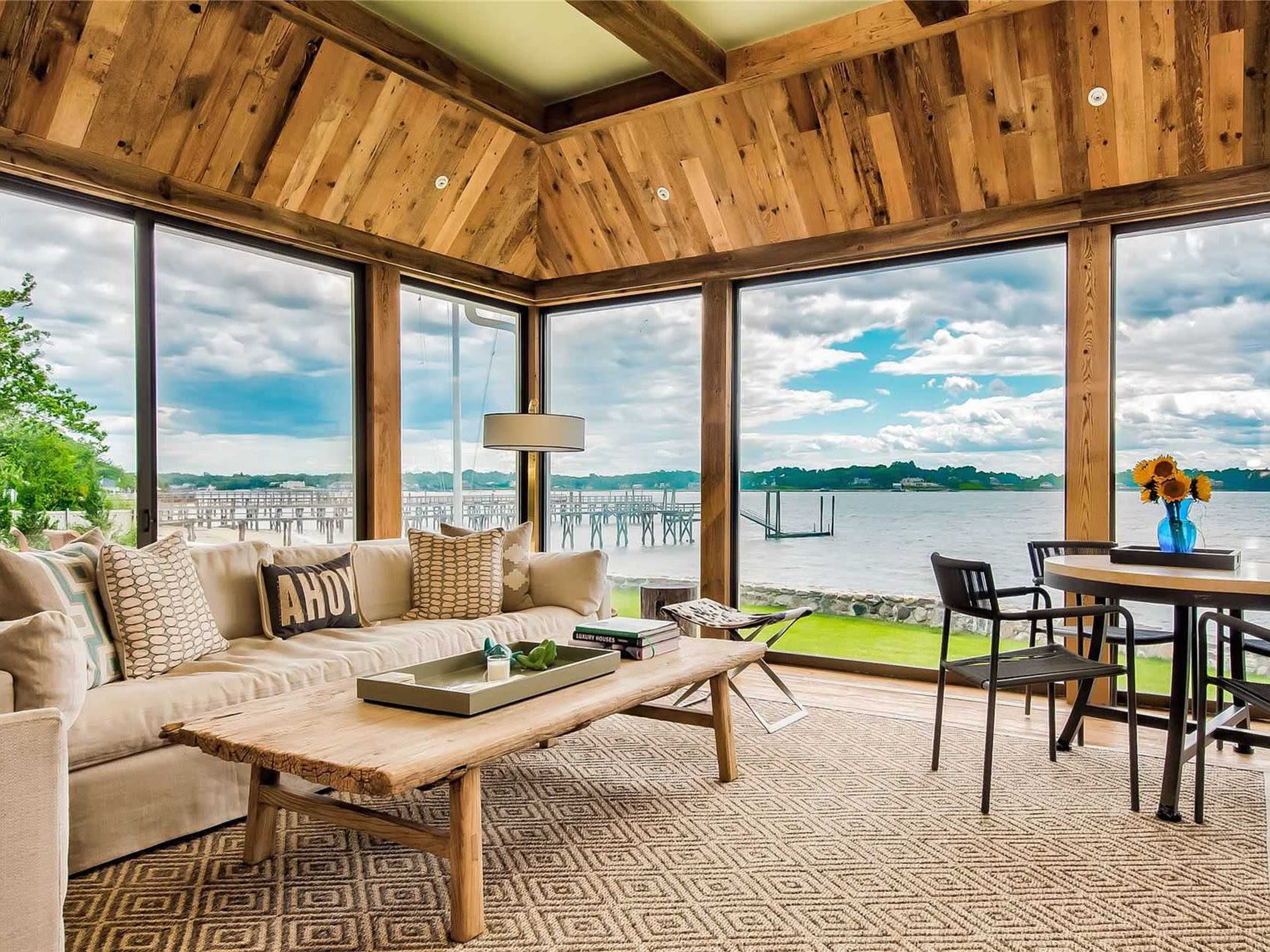 The image shows a cozy living area with large windows overlooking a waterfront view, featuring a wooden coffee table and a dining table with chairs.