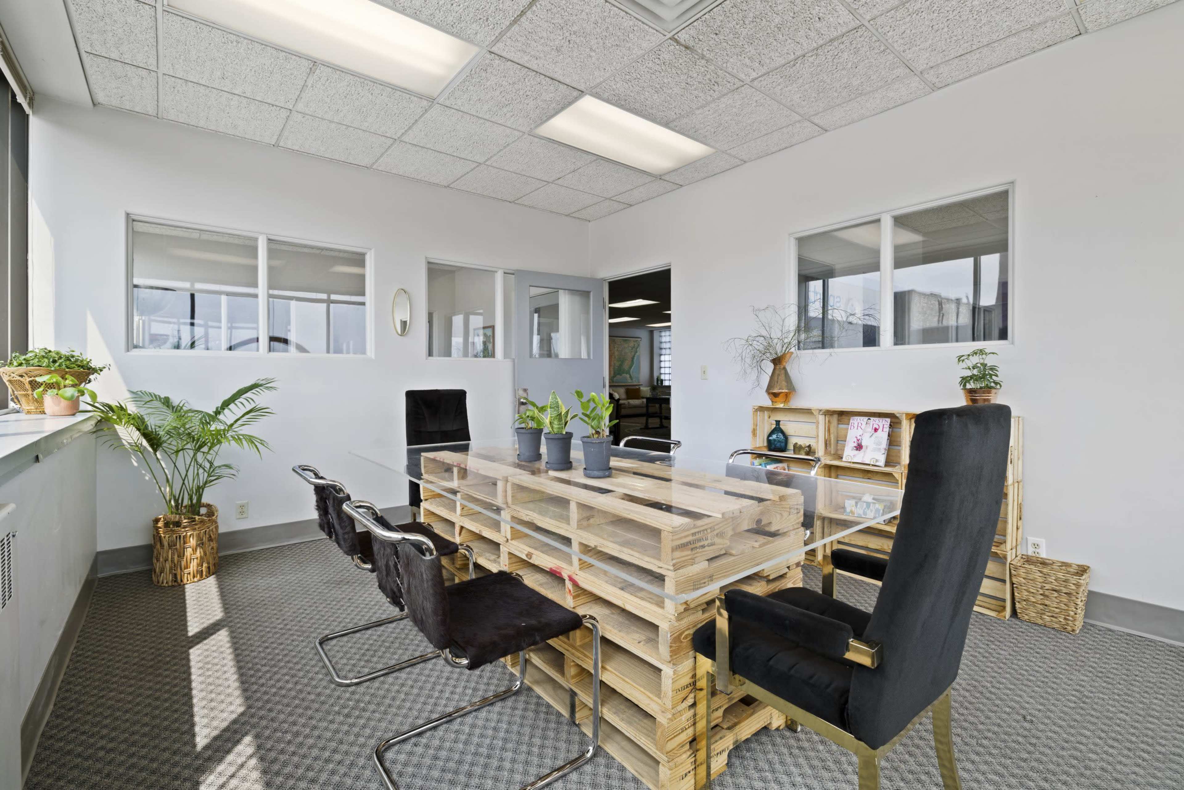 A meeting room features a large table made of wooden pallets surrounded by black chairs and plants in the corners.