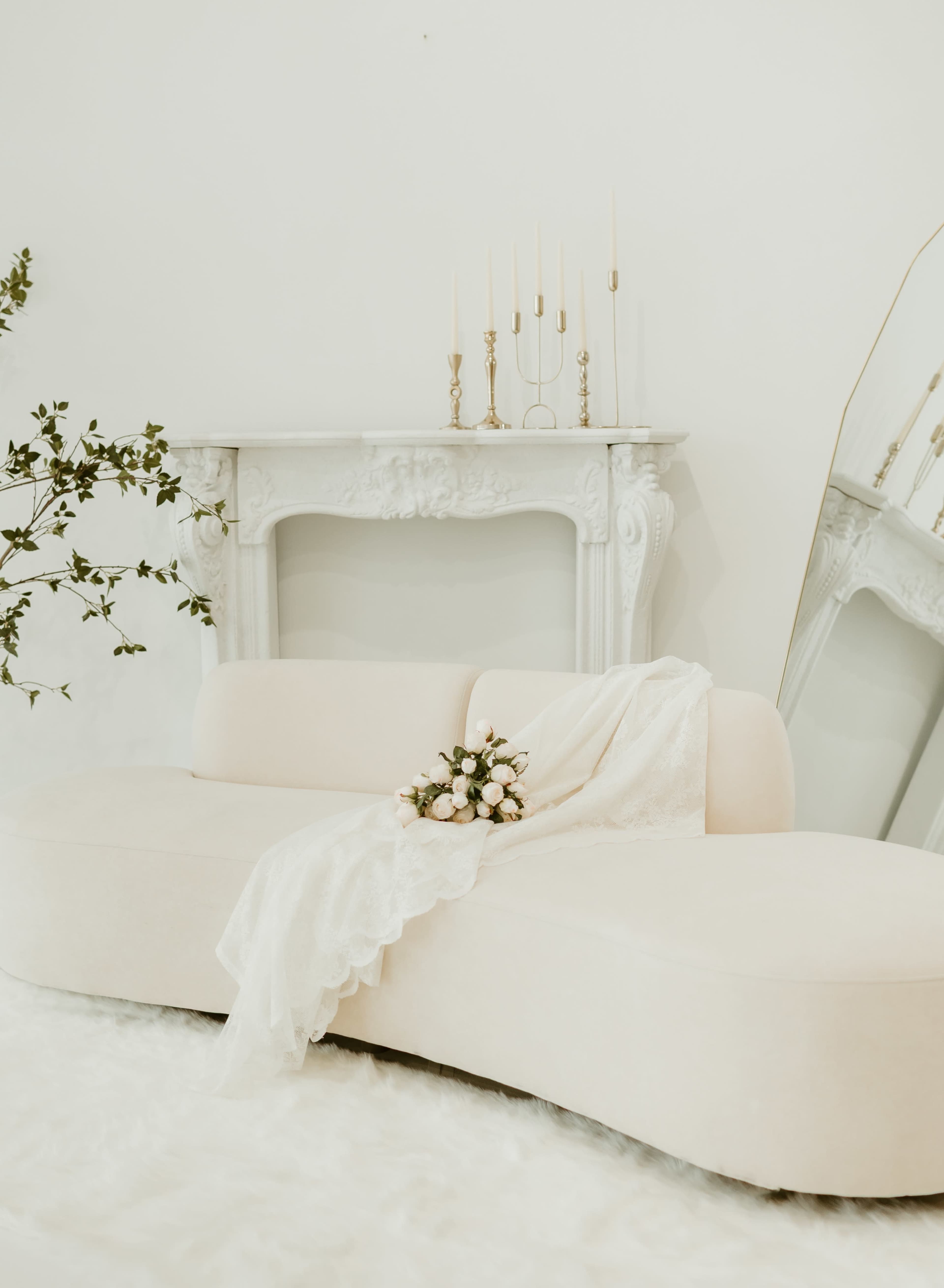 A light-colored sofa is positioned in front of a decorative white mantel, with a bouquet resting on the seat and a lace throw draped over it.