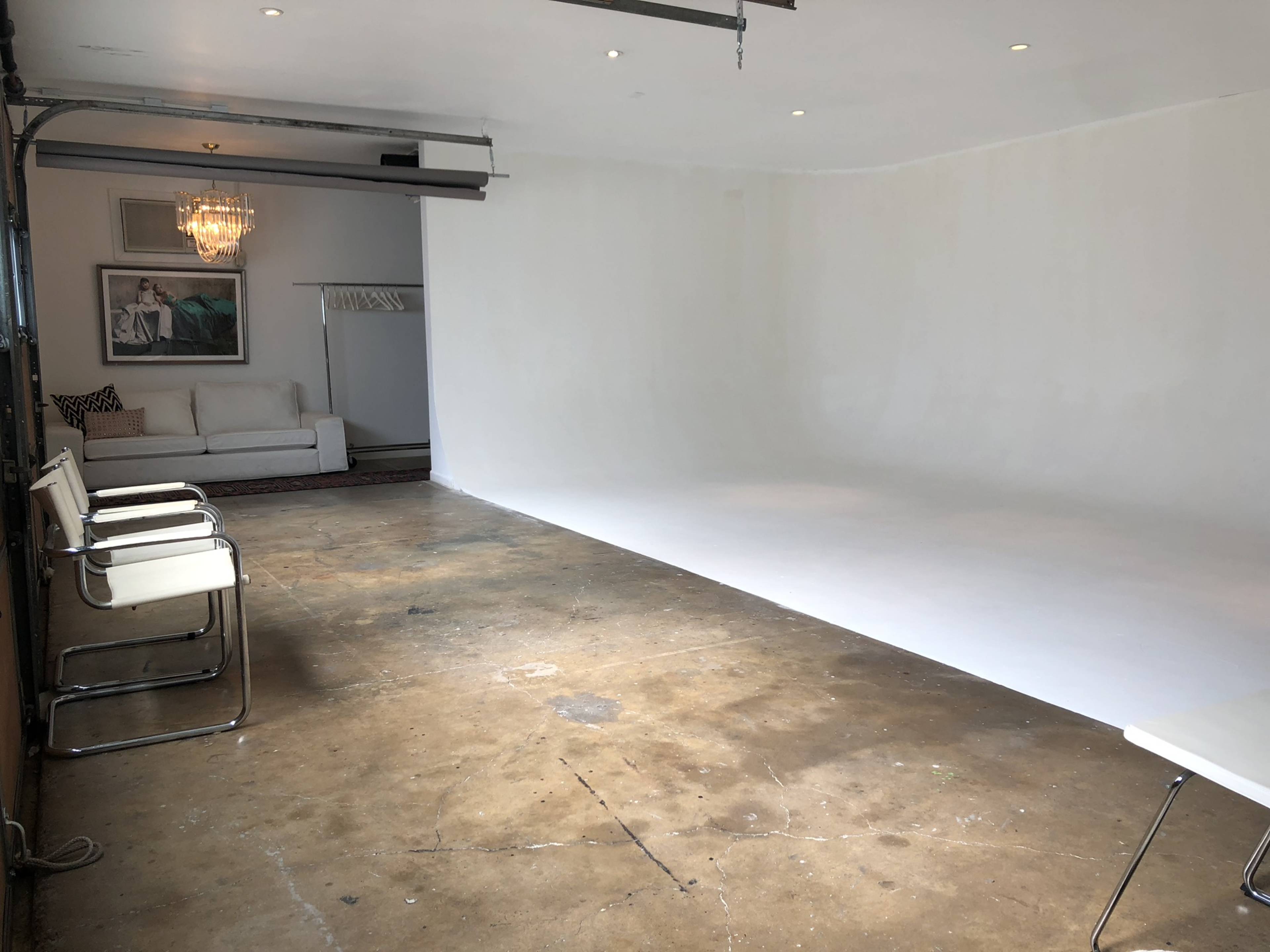 A spacious, empty studio with a white backdrop, polished concrete flooring, a sofa, and a decorative chandelier.