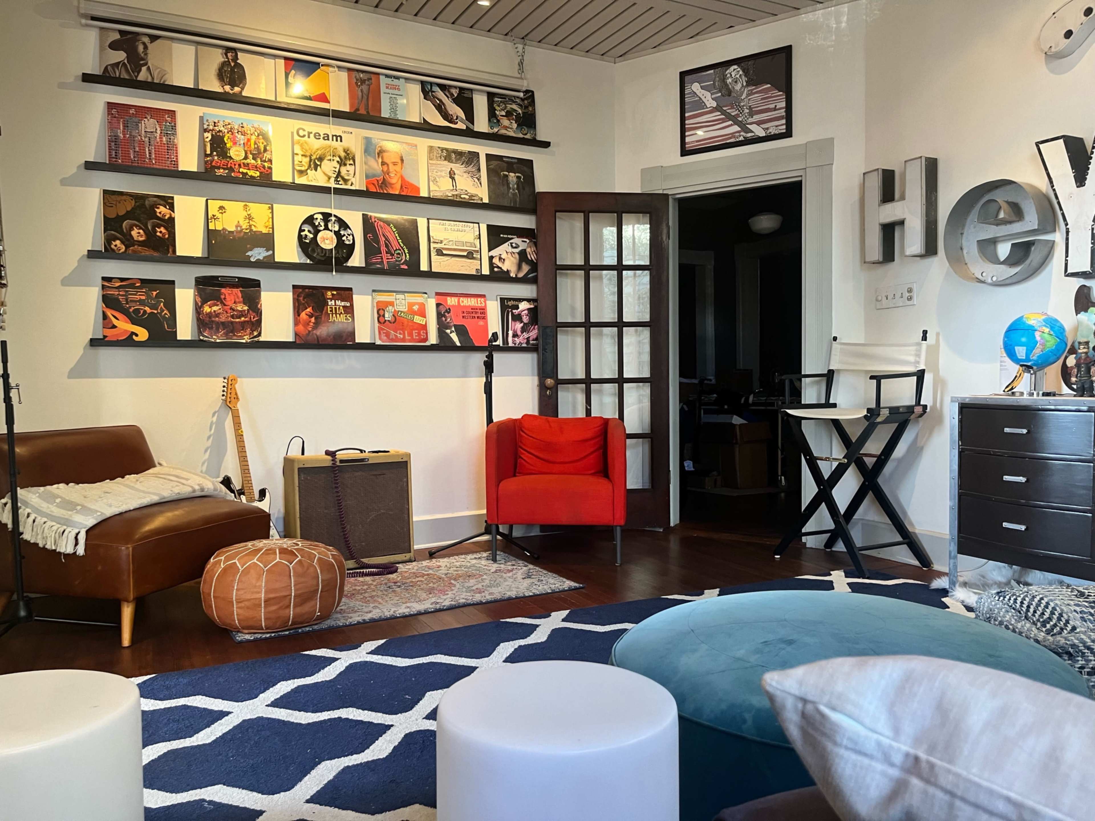 A cozy room with a wall displaying framed music albums, a brown leather couch, a red armchair, and a variety of seating options on a patterned rug.